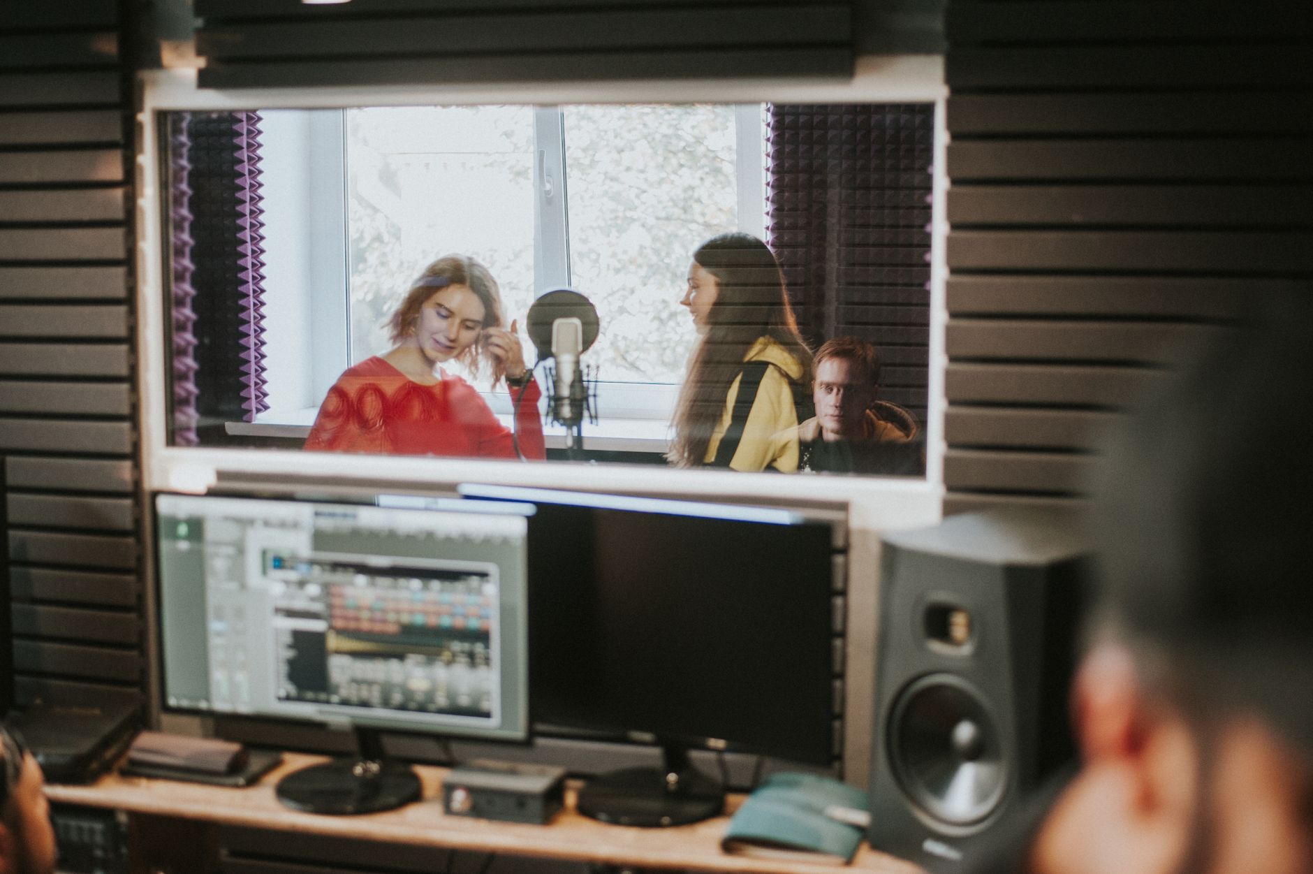 Artists recording music in a professional studio setting, captured through a glass window.
