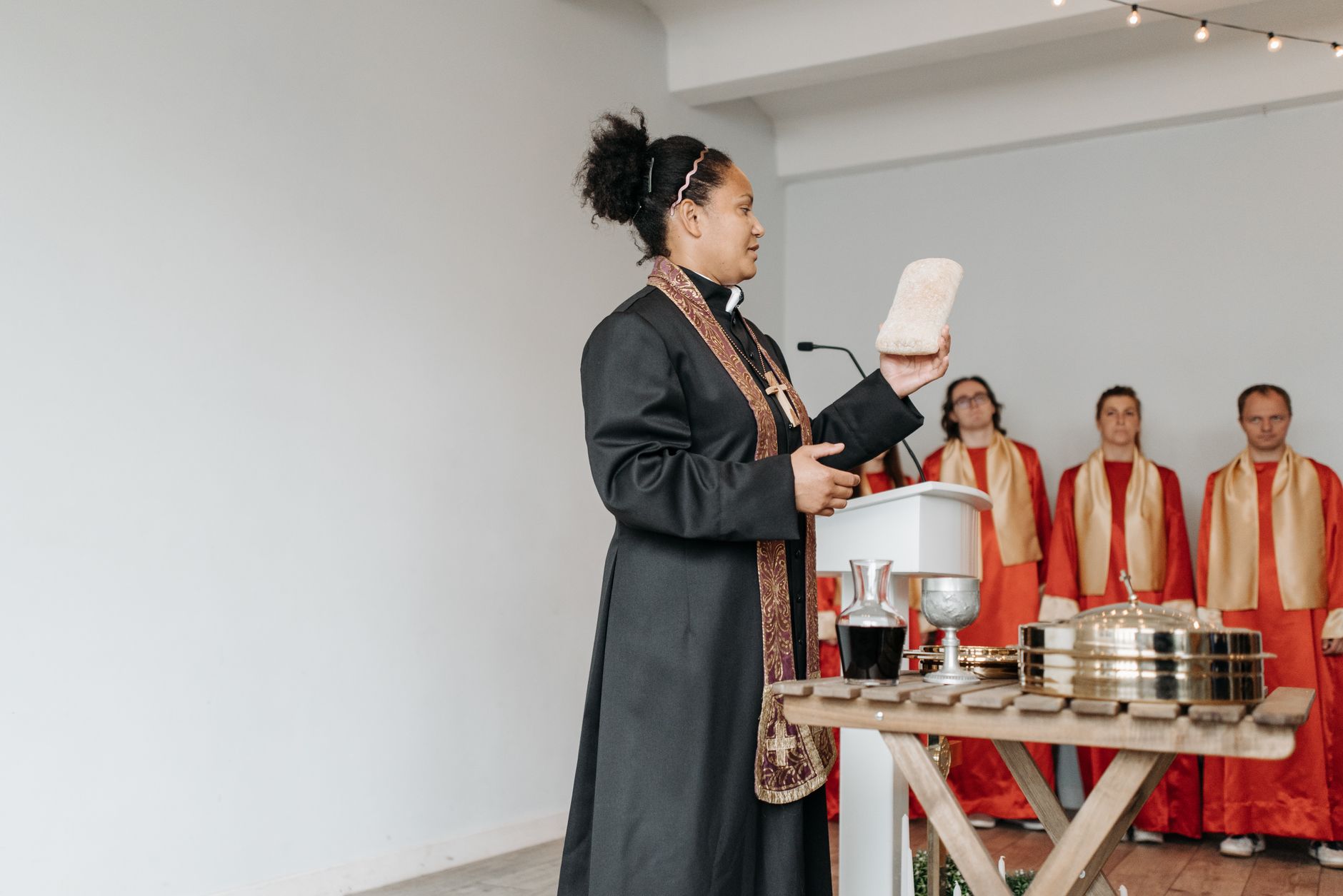 A female pastor conducts a communion service indoors with choir members in background.