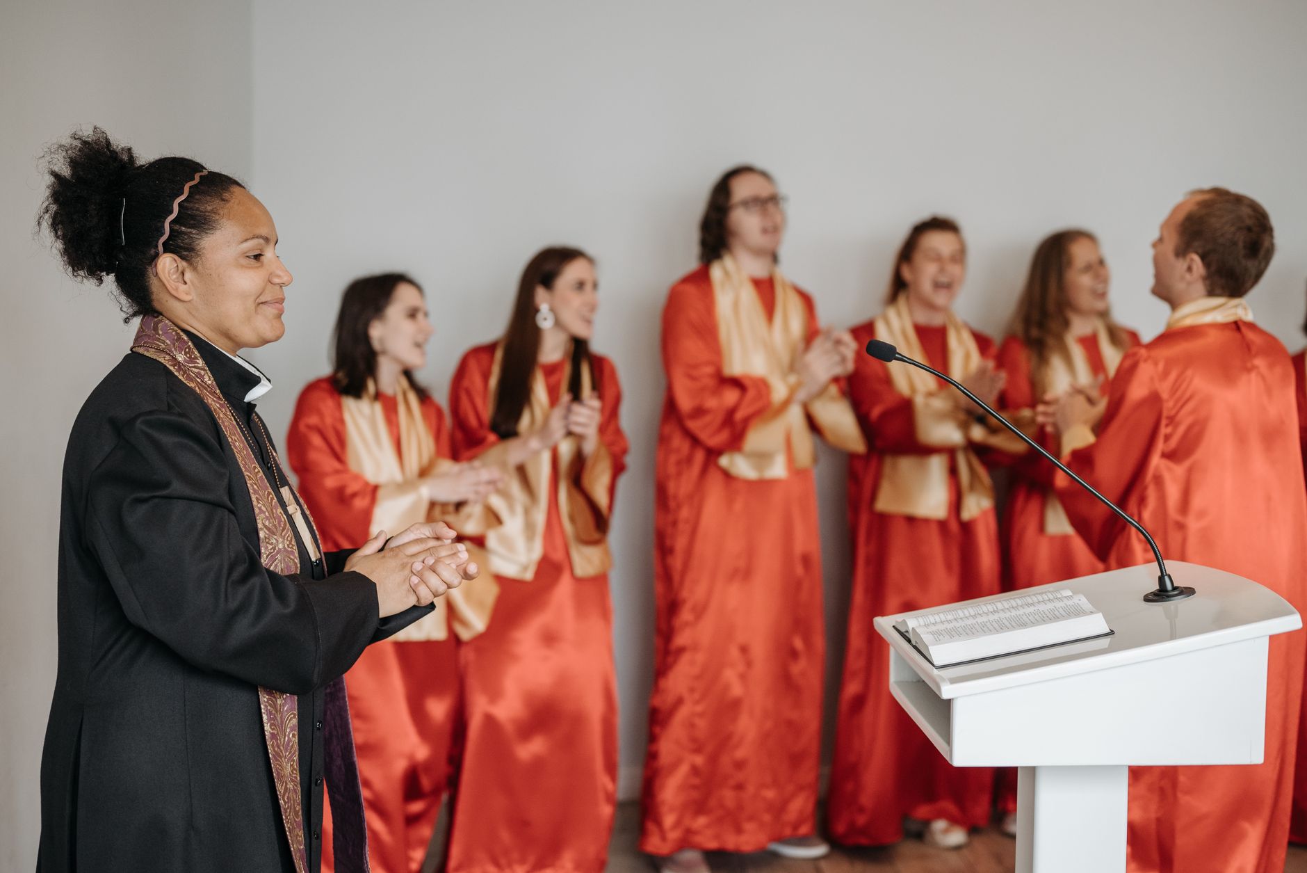A diverse choir claps and sings passionately in a church setting led by a woman pastor.