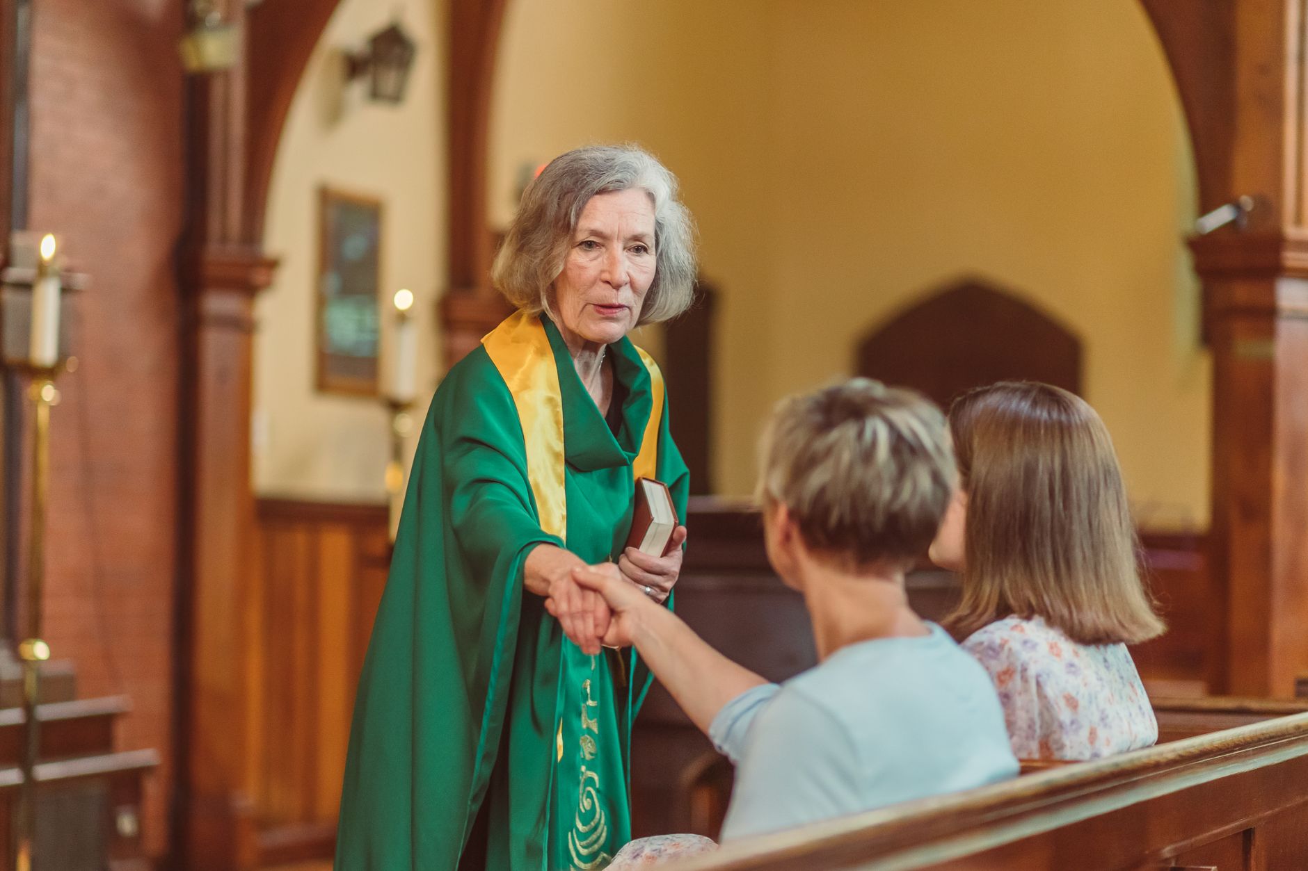 Female pastor in green robe warmly greeting congregants inside a church.