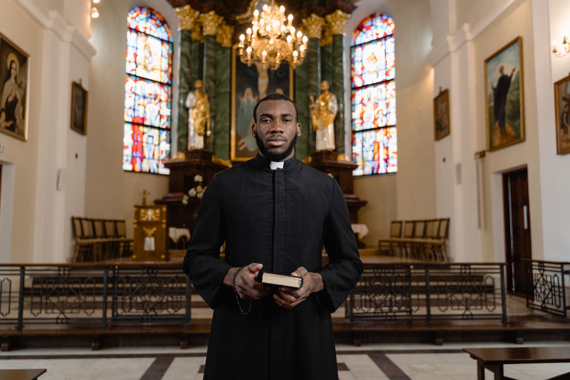 A priest stands in a church holding a Bible, surrounded by stained glass.