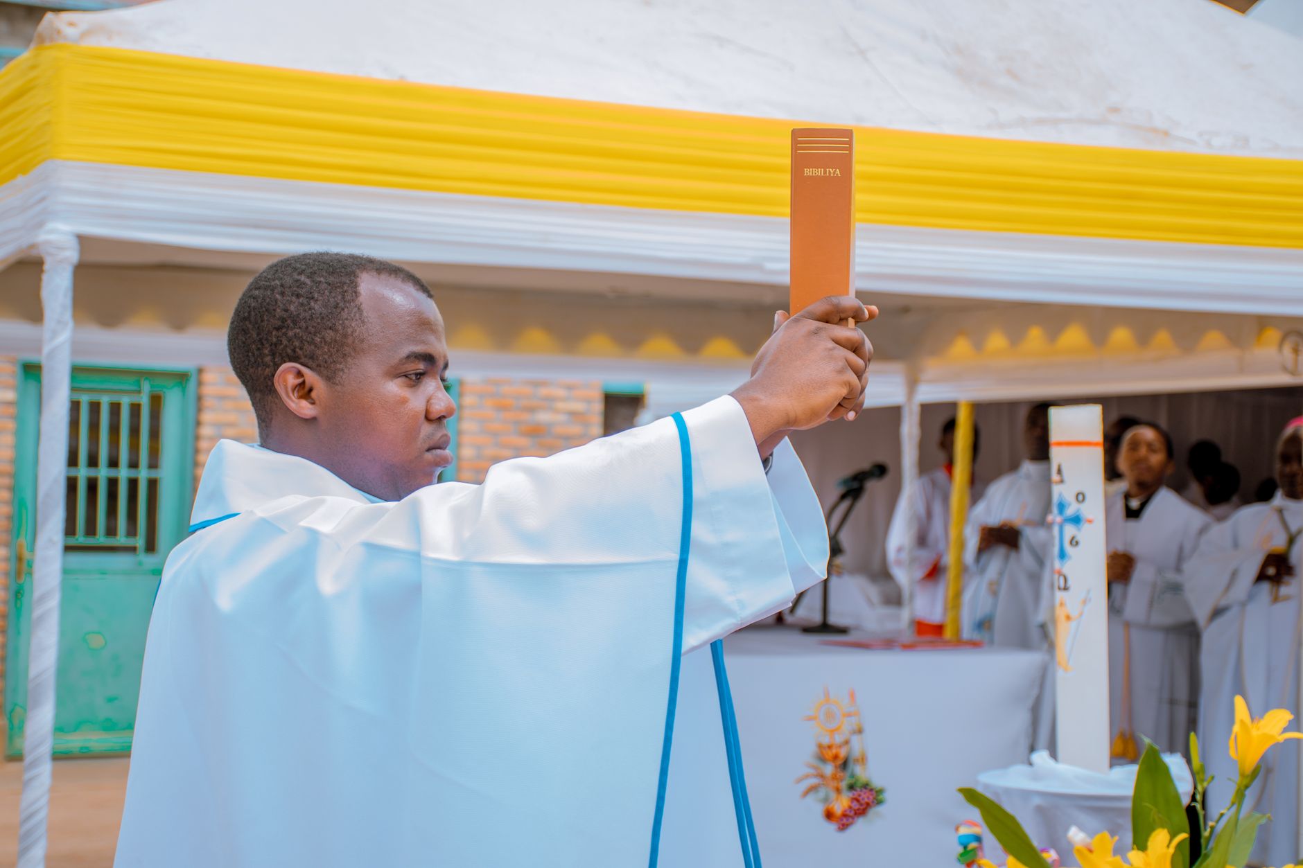 Priest in white robe elevates a Bible during an outdoor religious ceremony.