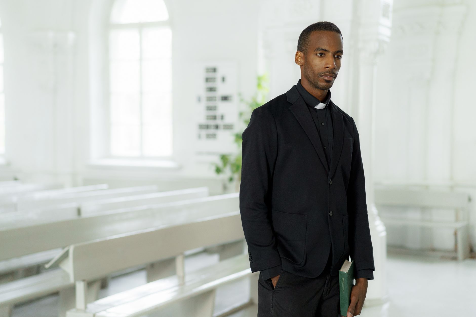 A pastor stands thoughtfully in a bright church, holding a book with confidence.
