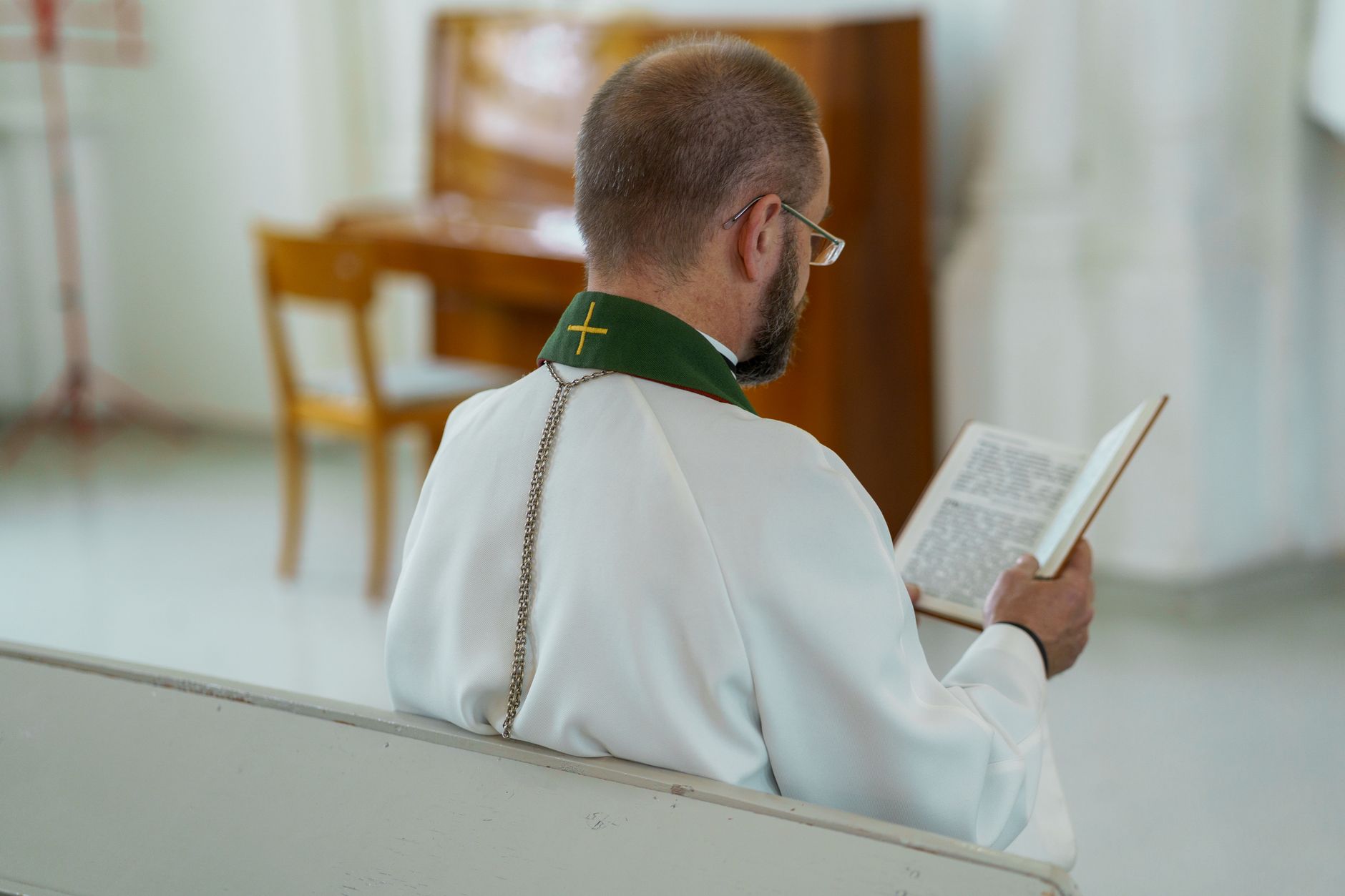 A priest in a white robe reads a holy book while sitting in a church pew.