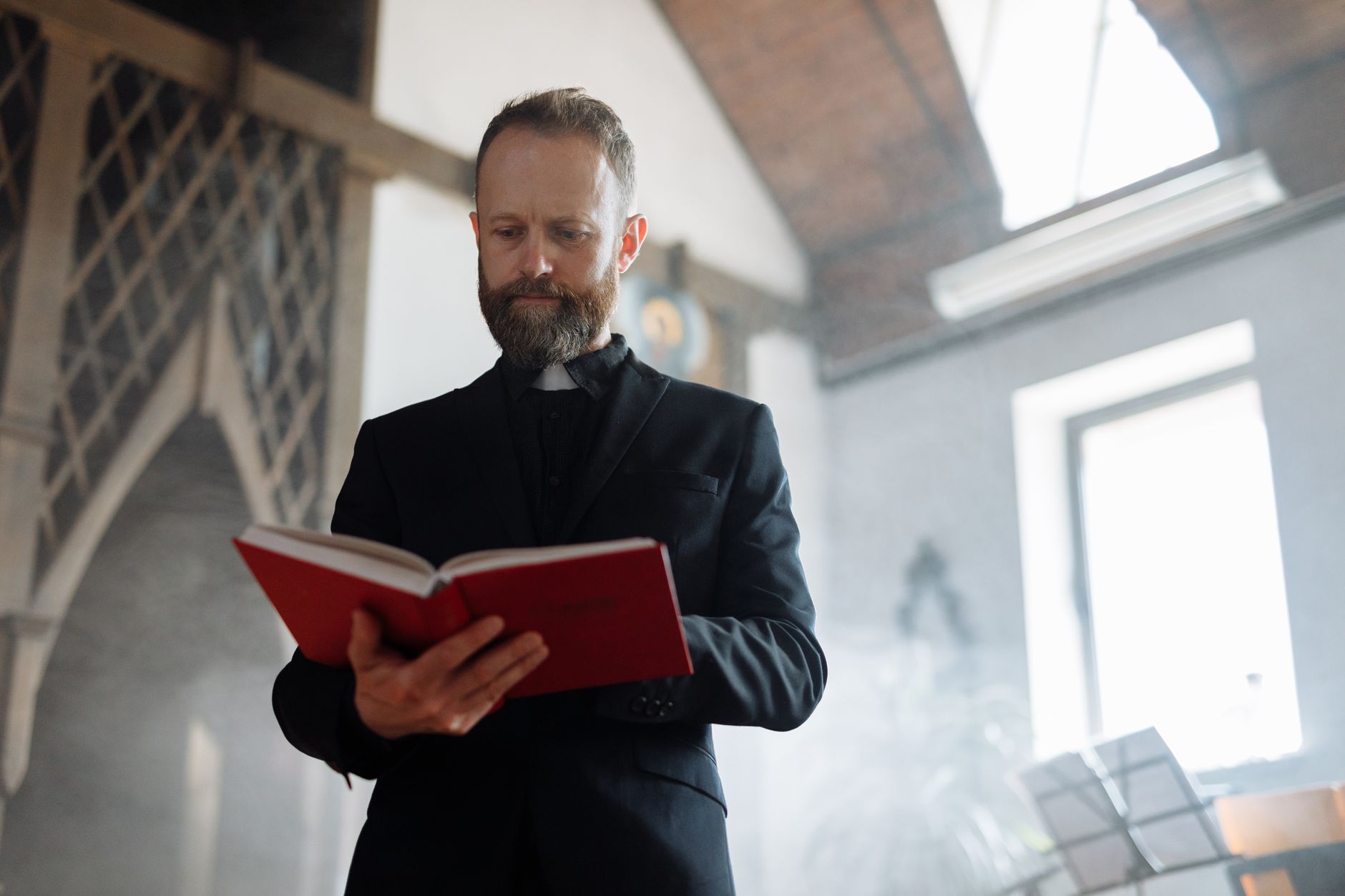 A Catholic priest stands in a church, reading a red book during the day.