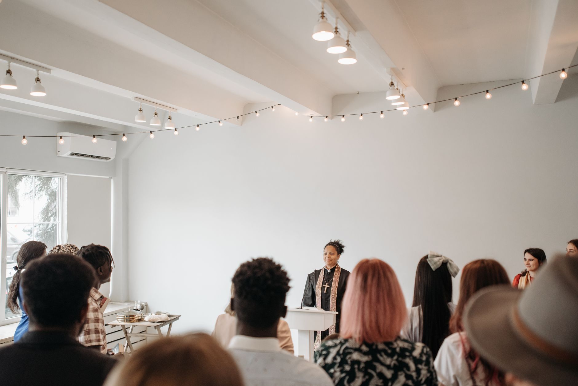 A diverse group of people listens to a pastor in a well-lit church with string lights.