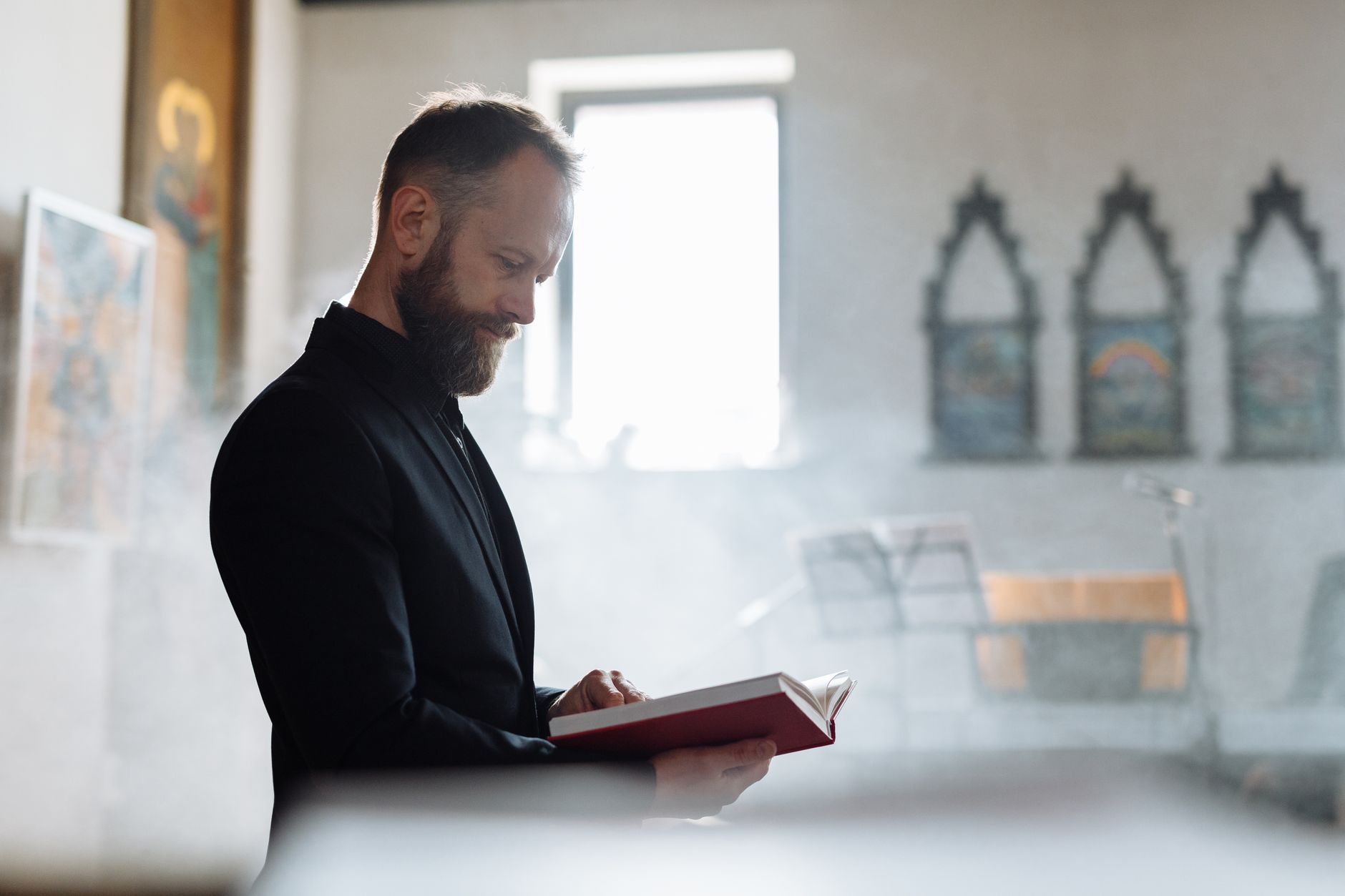 Priest in cassock reading a bible in a church with soft light and smoke.