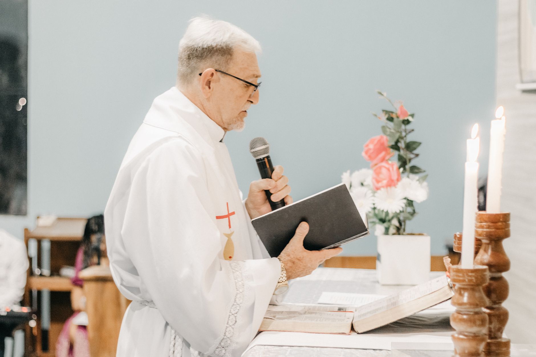 Side view of a Catholic priest in vestments, holding a microphone and book, conducting a ceremony.