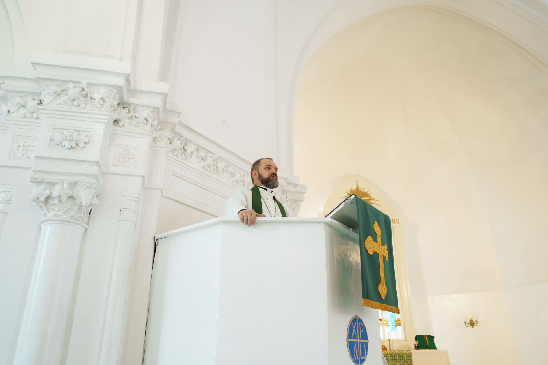 A priest in a white and gold ornate church delivering a sermon from the pulpit.