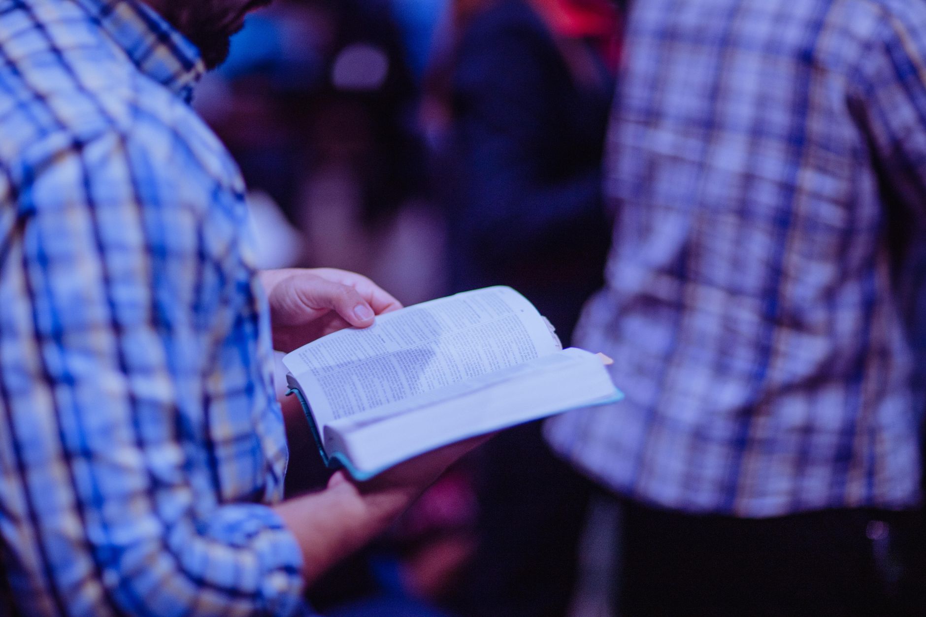 A man stands indoors reading a Bible, focusing amidst a crowd.