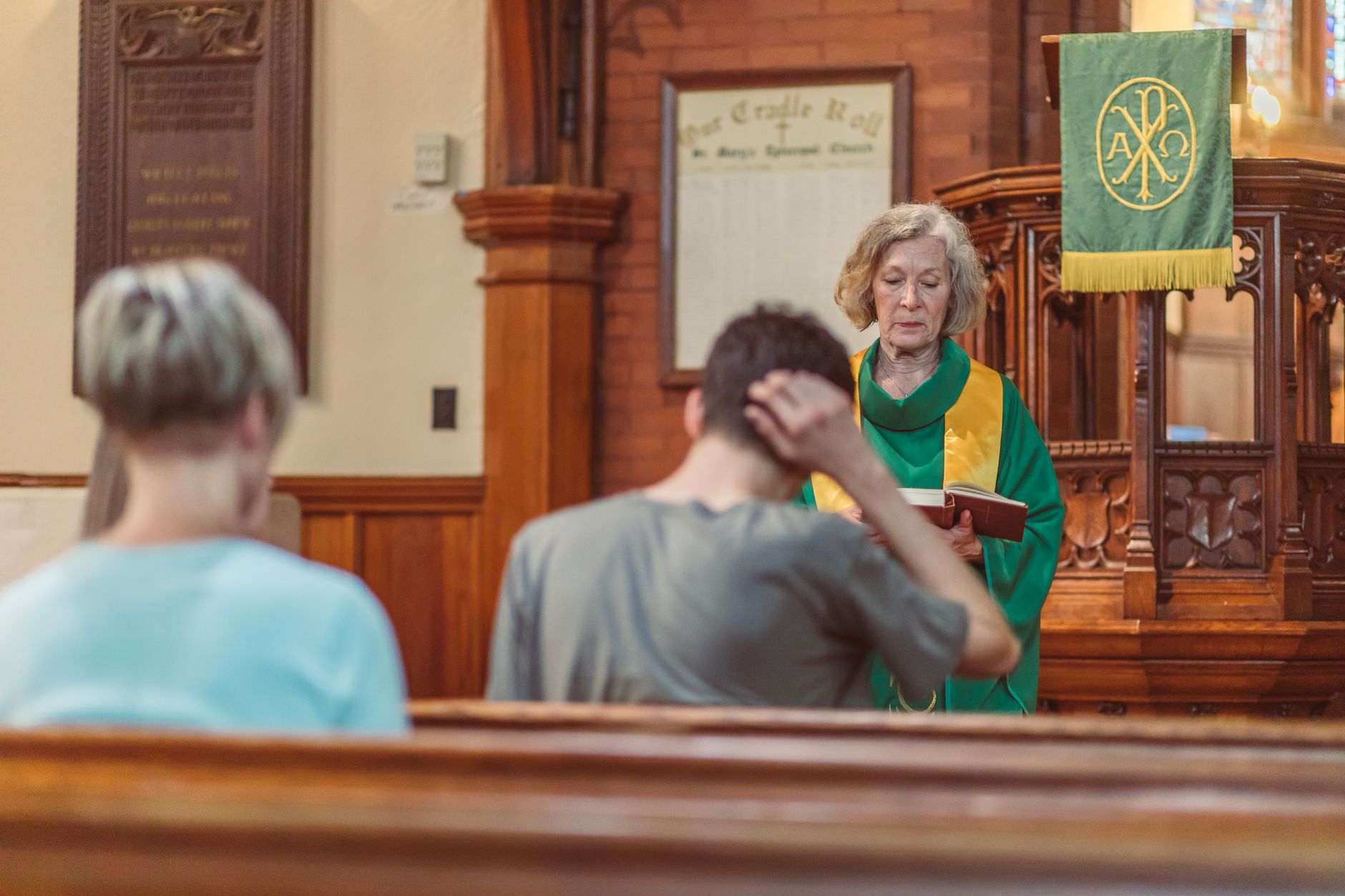 An elderly female clergyperson leads a service inside a church, engaging with the congregation.