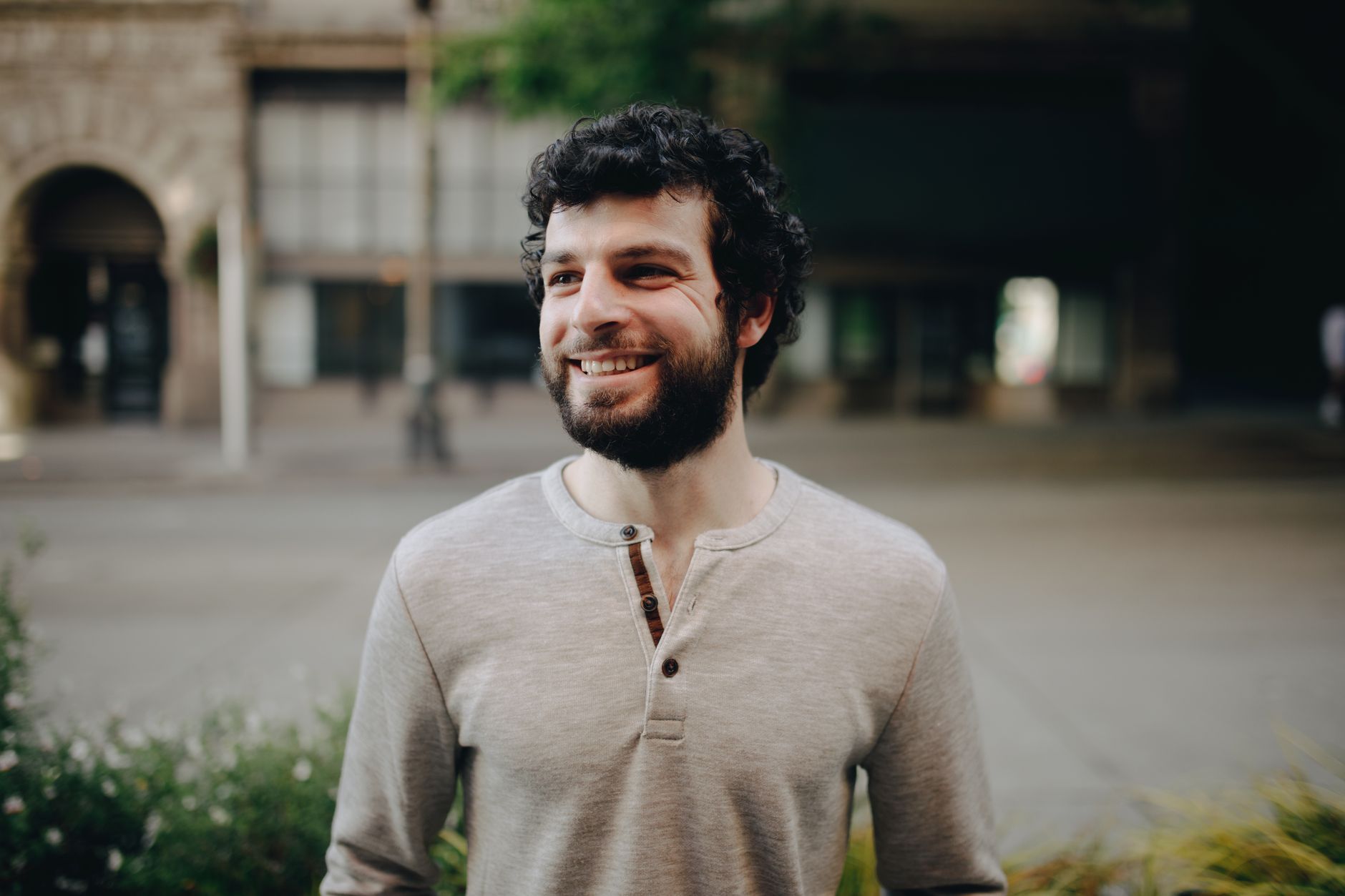 A bearded man with curly hair smiling outdoors in a casual setting.