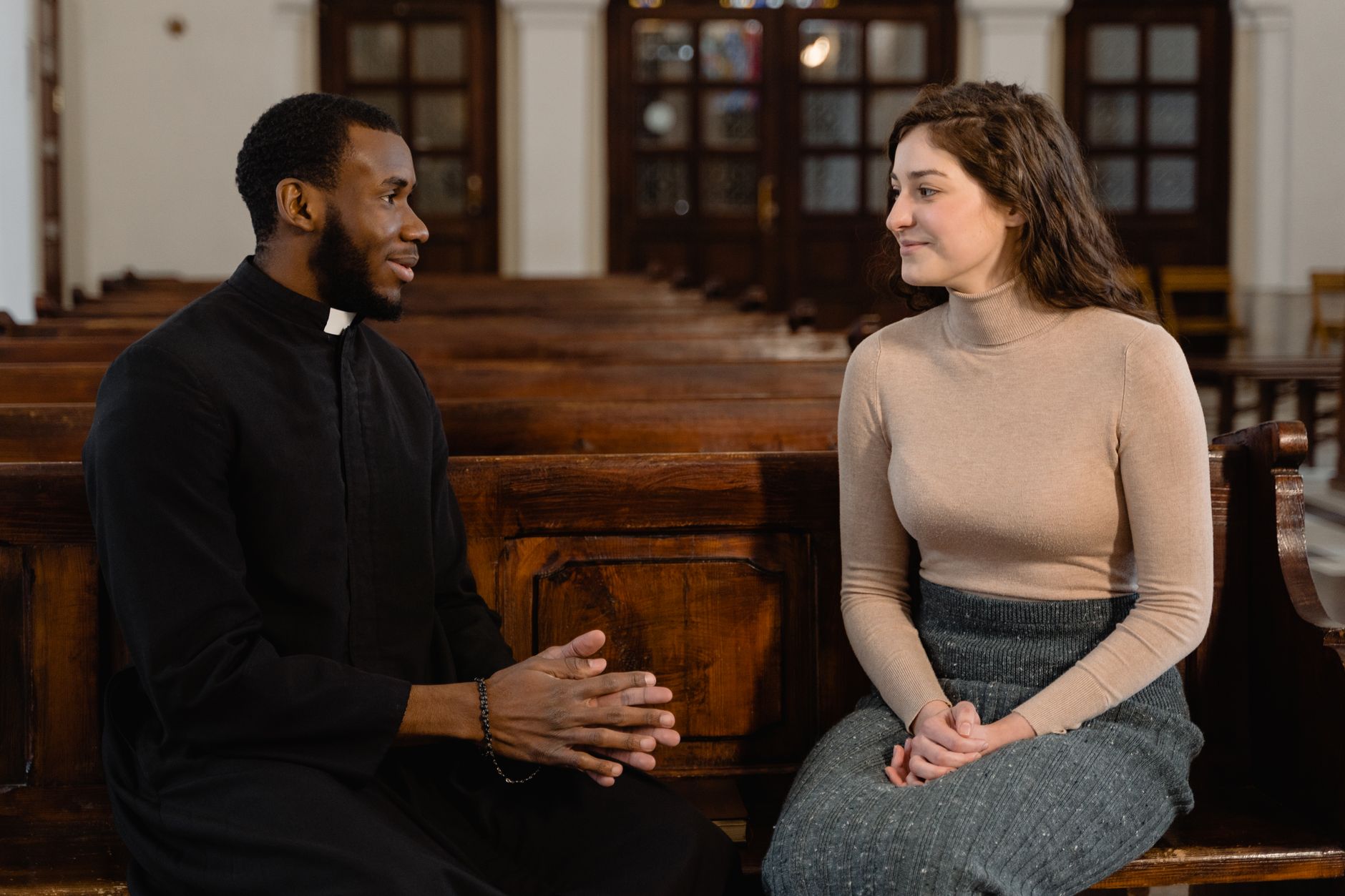 A priest and a woman seated on wooden pews in discussion, symbolizing faith and community.