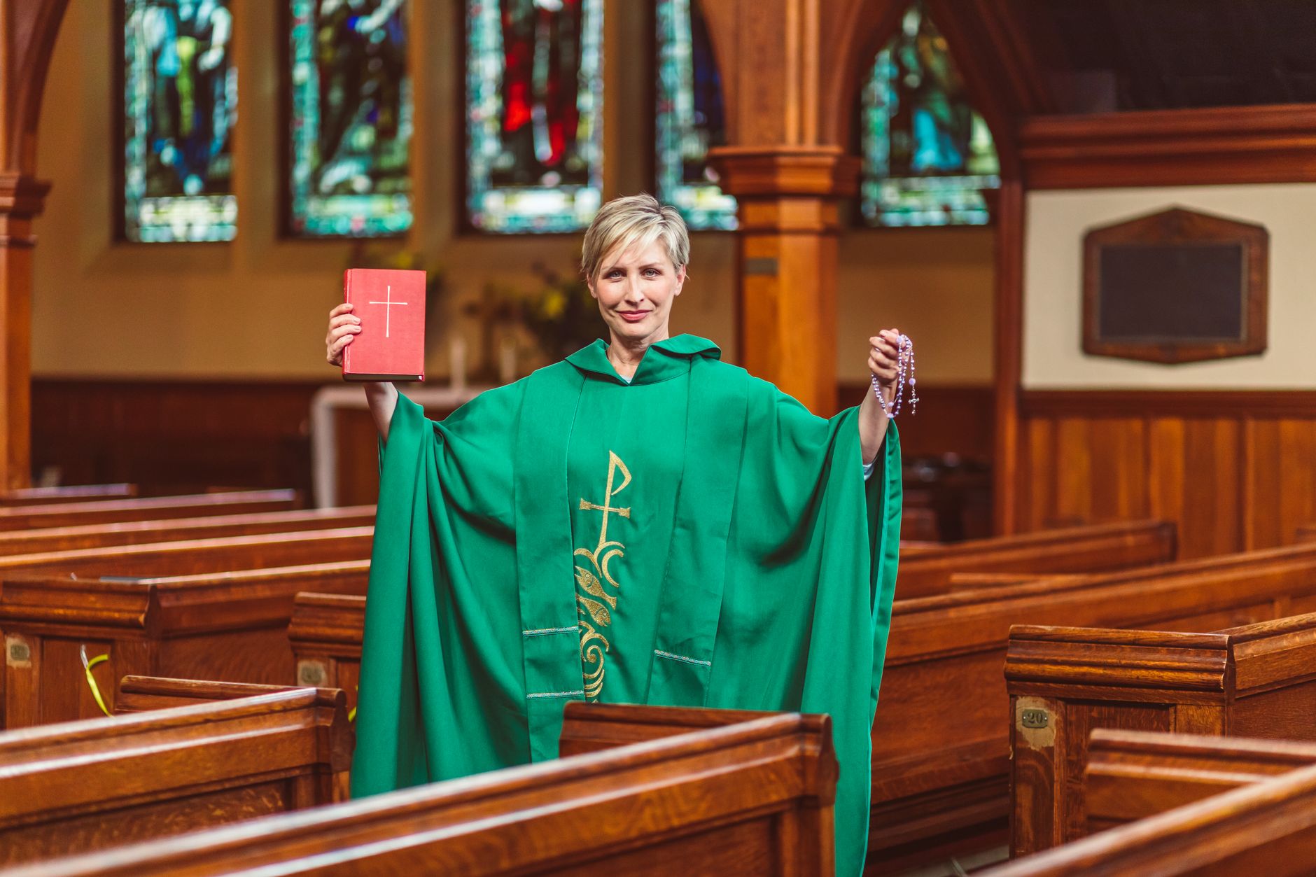 Caucasian female priest in green robe holding Bible and rosary inside a church with stained glass windows.