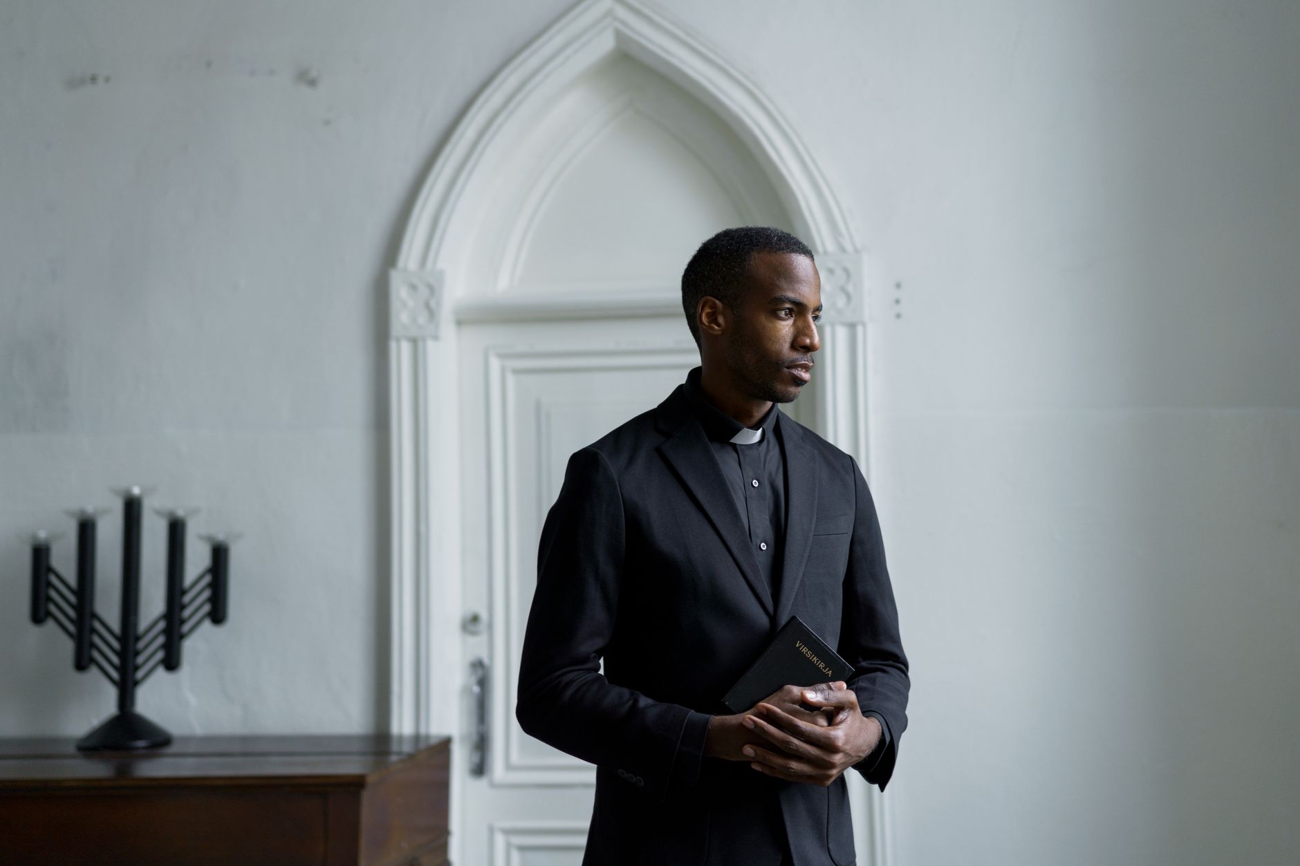 A thoughtful pastor stands indoors holding a Bible, showcasing serenity and faith.