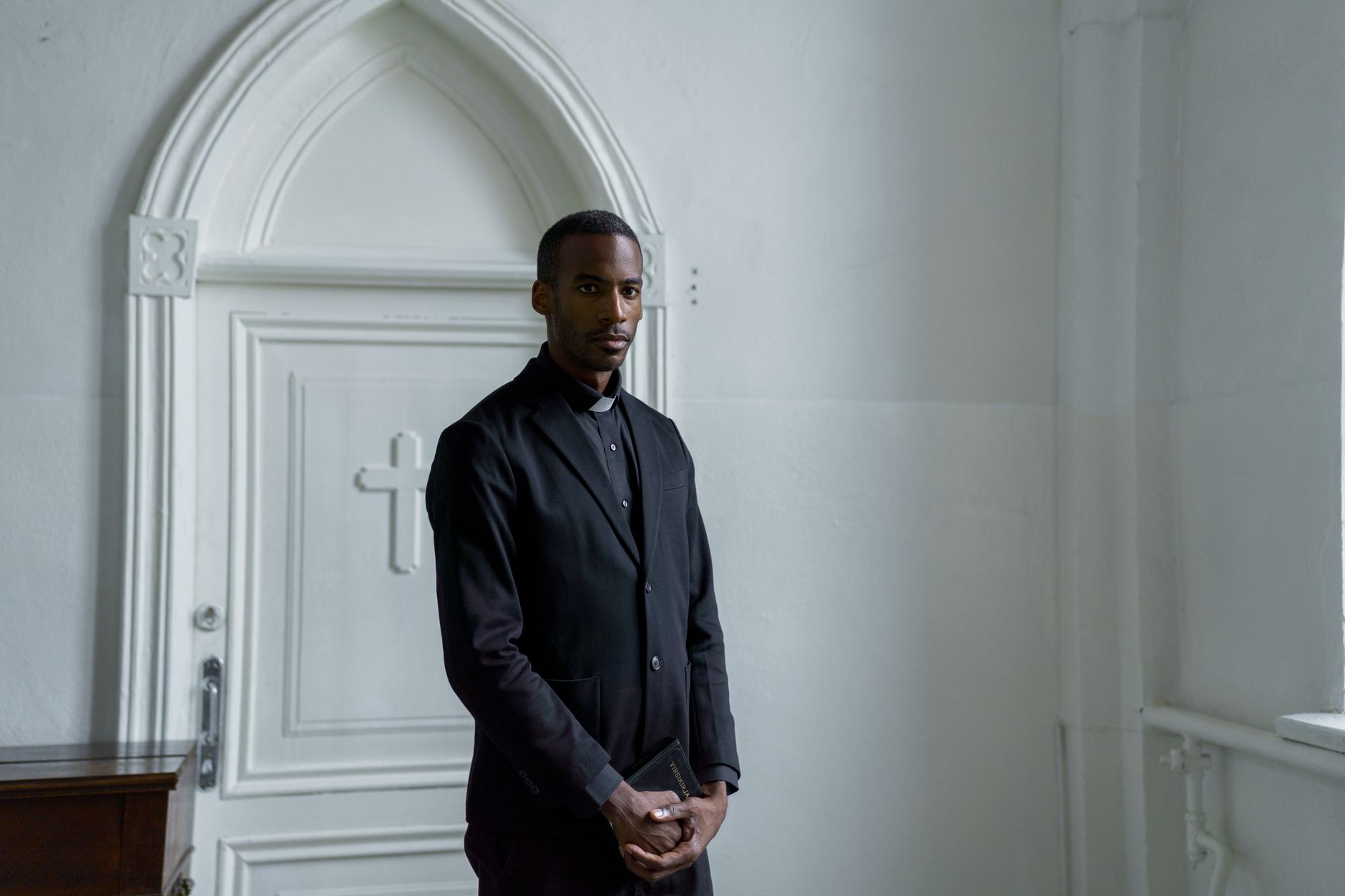 Young male minister stands confidently holding a book in front of a church door.