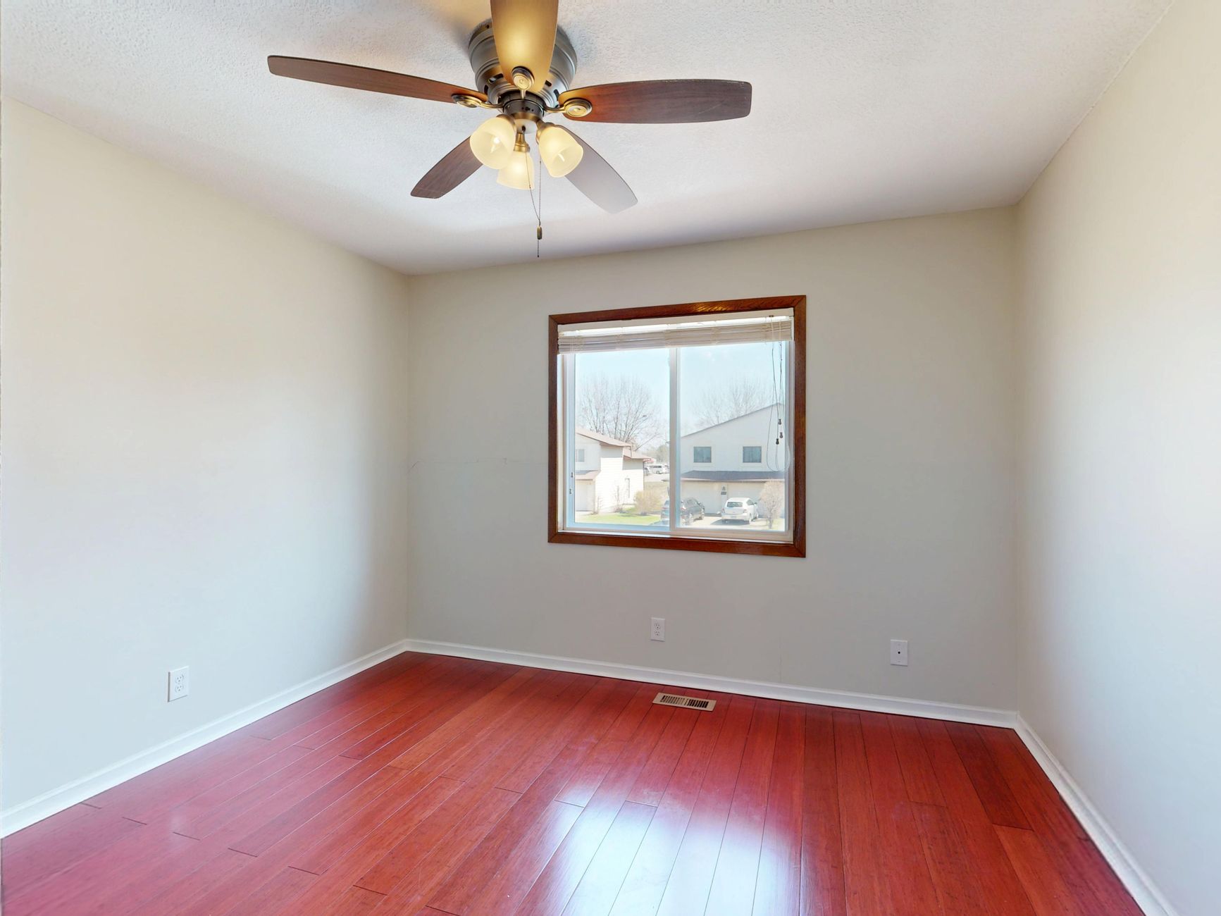 Spacious empty room with wooden flooring, window view, and ceiling fan.