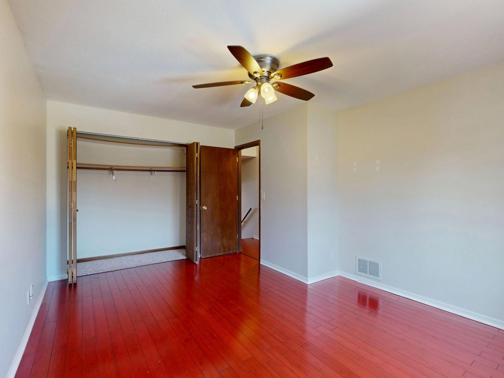 An empty room with polished wooden floors, a ceiling fan, and an open closet space.