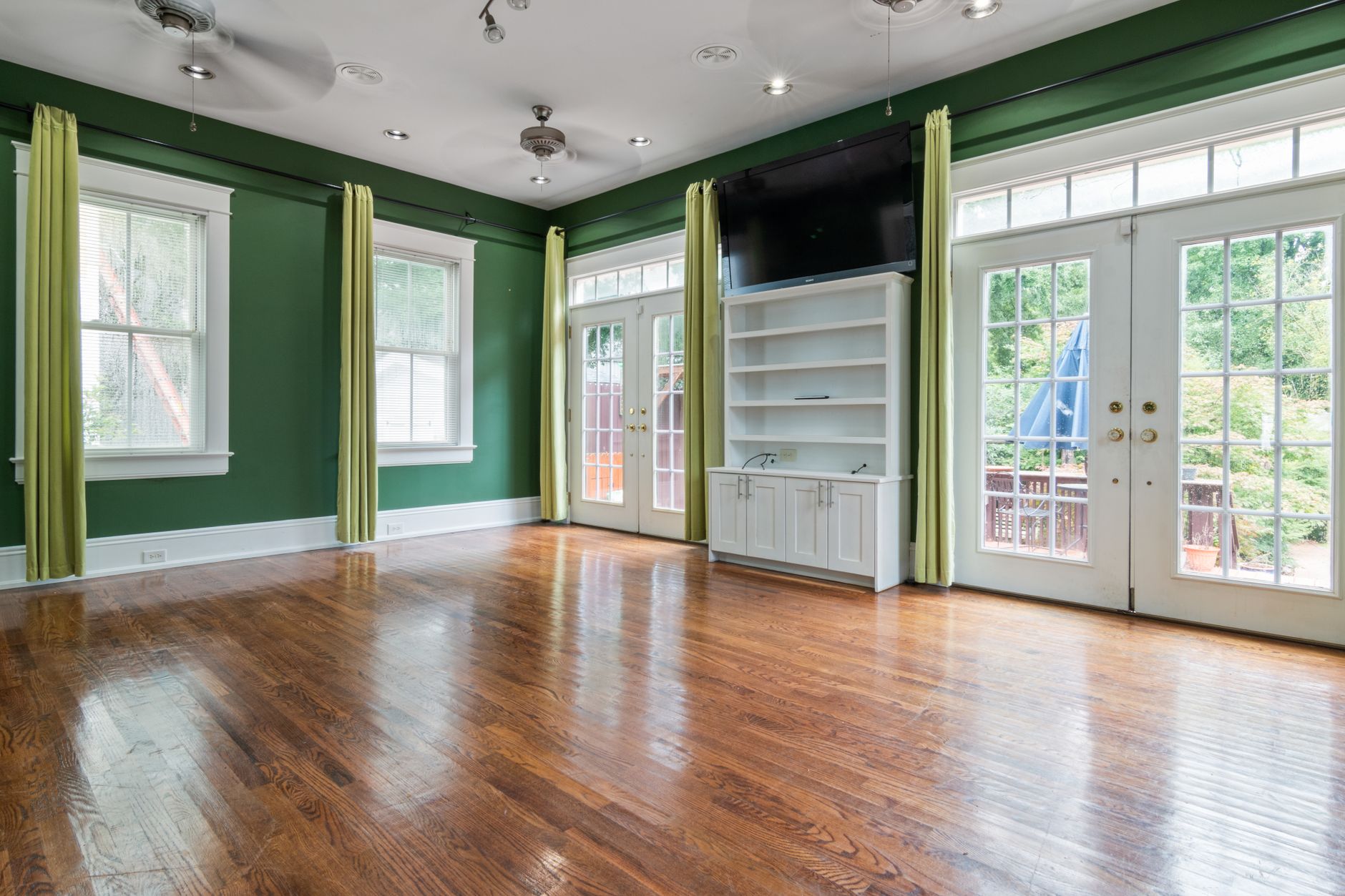 Empty living room with green walls, hardwood flooring, and large windows providing natural light.