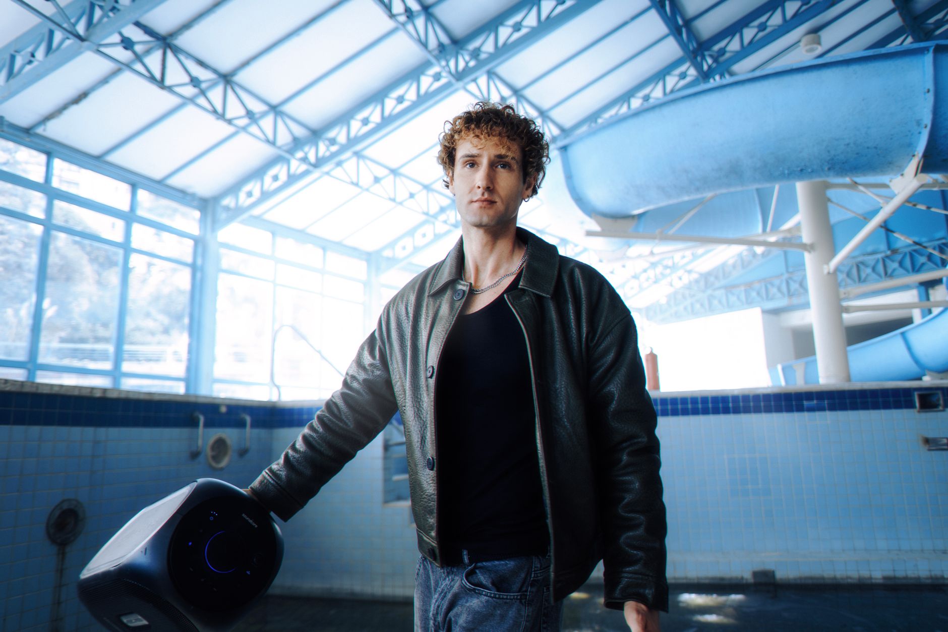 Man holding speaker in indoor pool setting with blue waterslide in background, wearing a leather jacket.