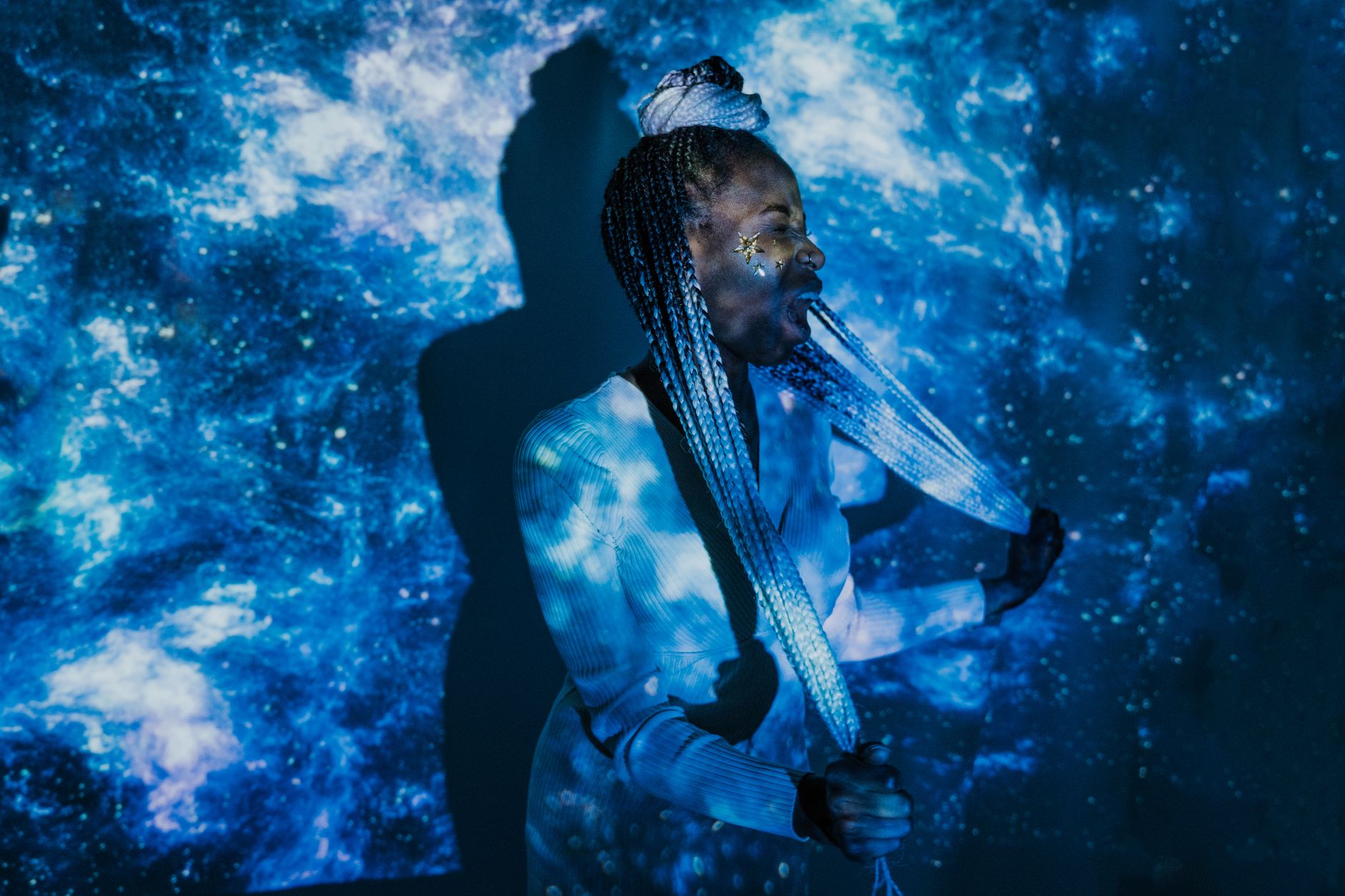 A stylish model with braided hair in an indoor setting featuring space-themed projections, creating a conceptual and artistic scene.