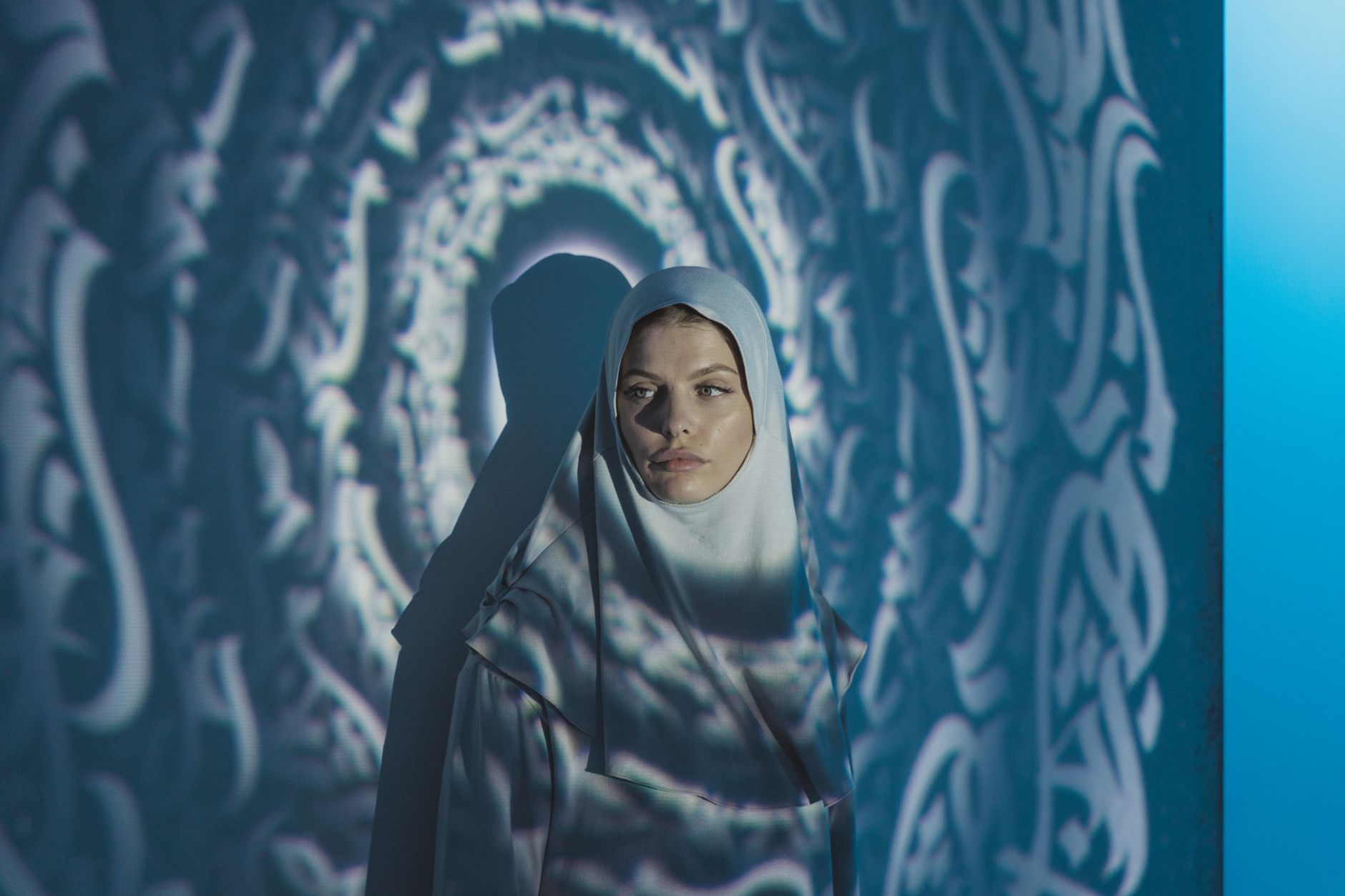 A woman in a hijab stands gracefully with shadow patterns projected around her, creating an elegant and artistic scene.
