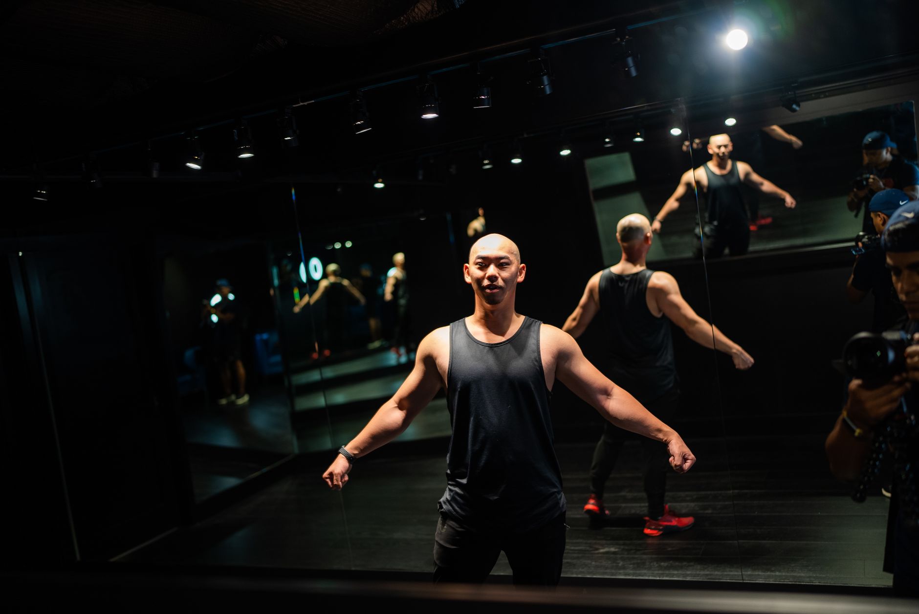 Bald, muscular man poses in a gym with mirror reflections wearing a tank top.