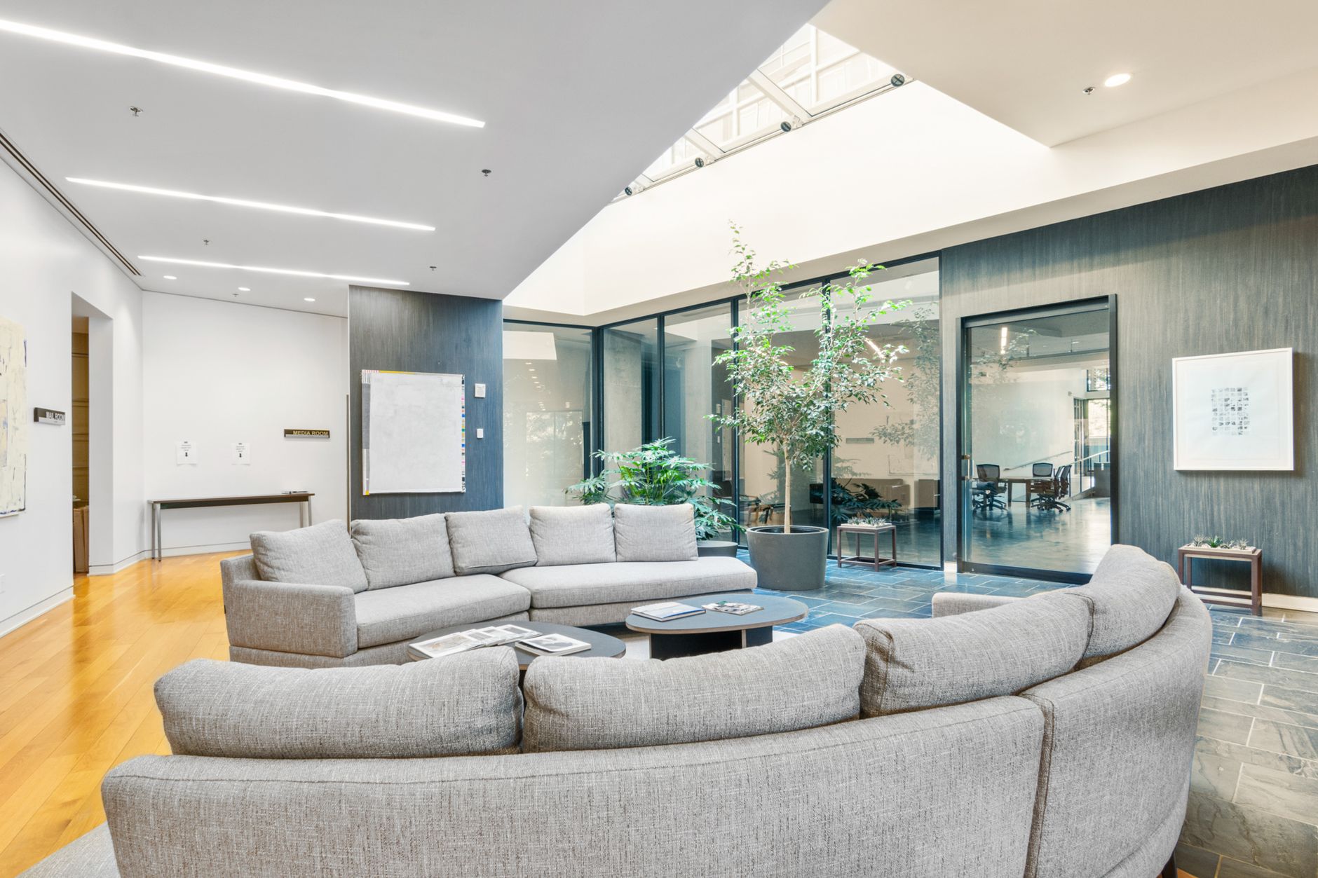 Spacious modern lounge area with grey sofa, glass walls, and indoor plants.