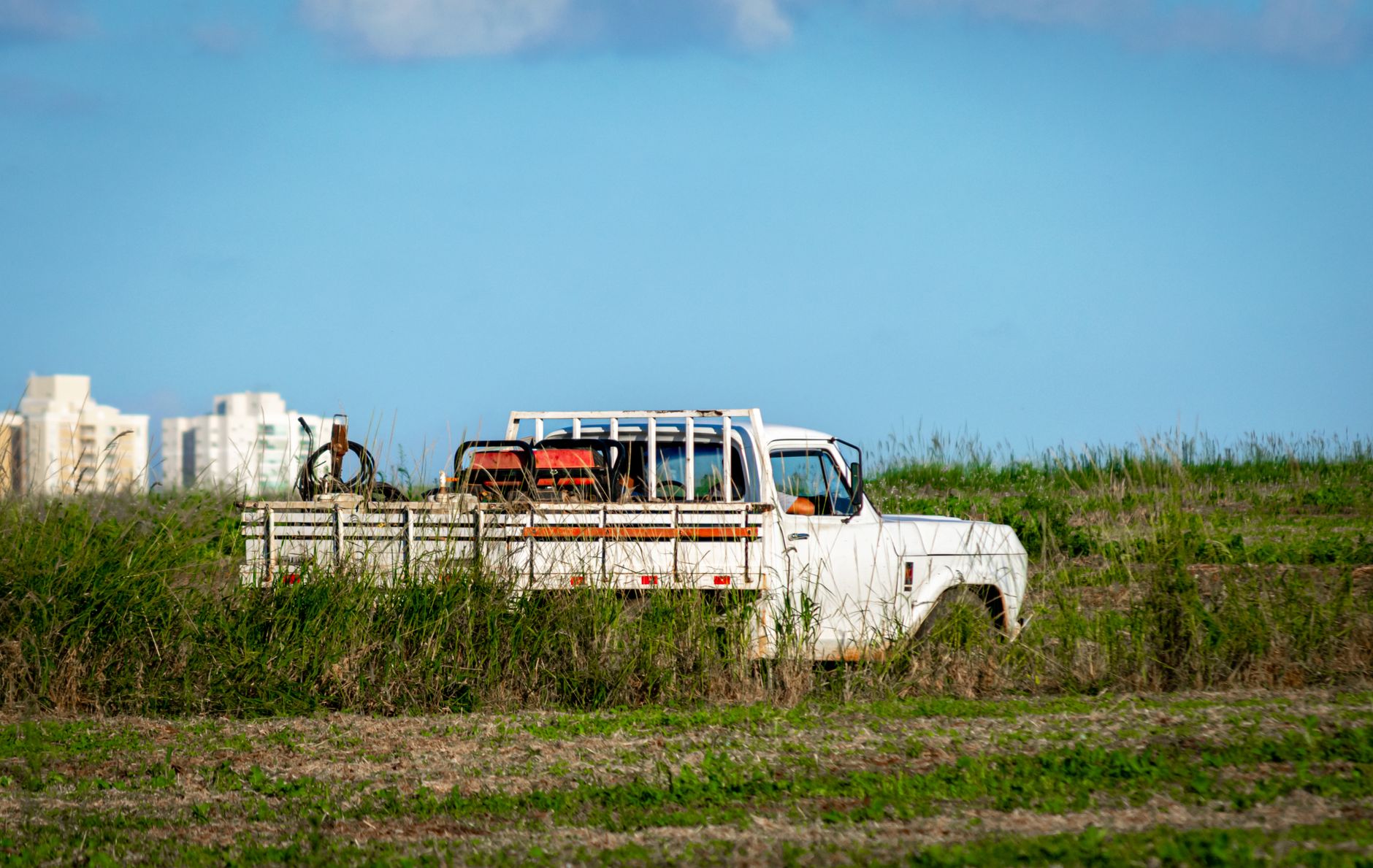 A rustic white pickup truck sits in a vast rural field with distant buildings framed by blue sky.