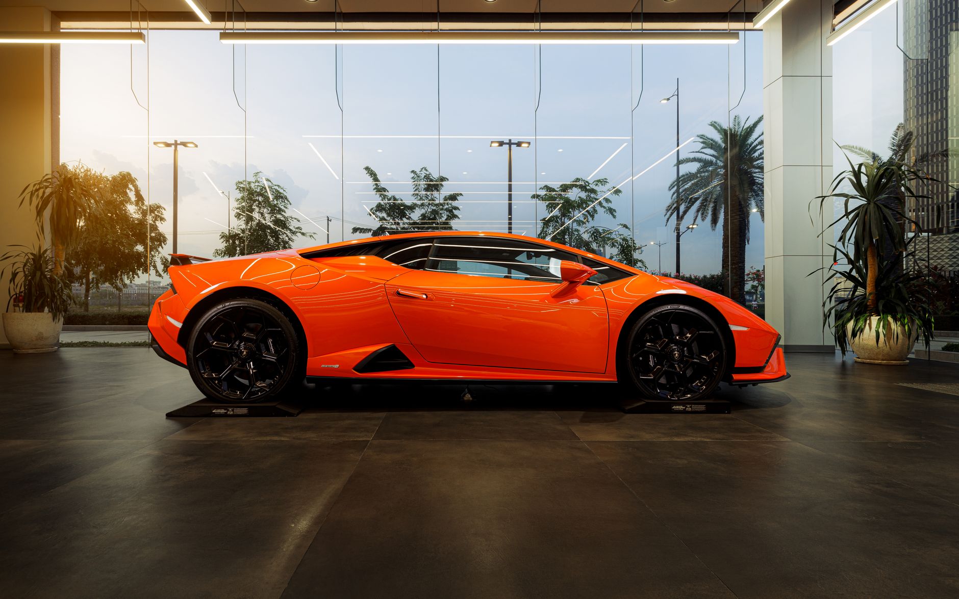 Profile view of a bright orange luxury sports car inside a modern showroom with a scenic outside view.