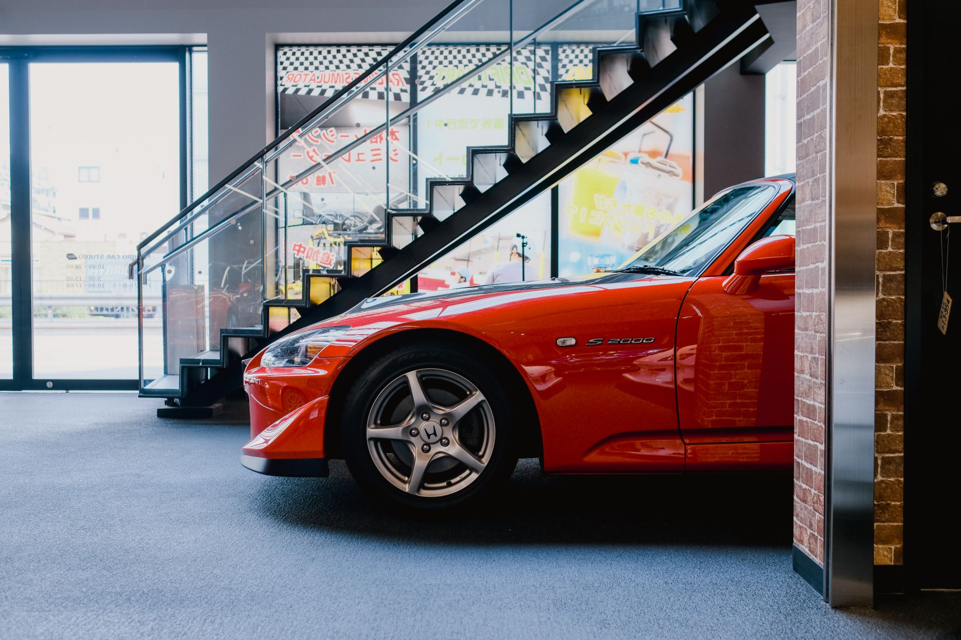 A sleek red Honda S2000 displayed indoors in a contemporary car showroom with stylish glass staircase.