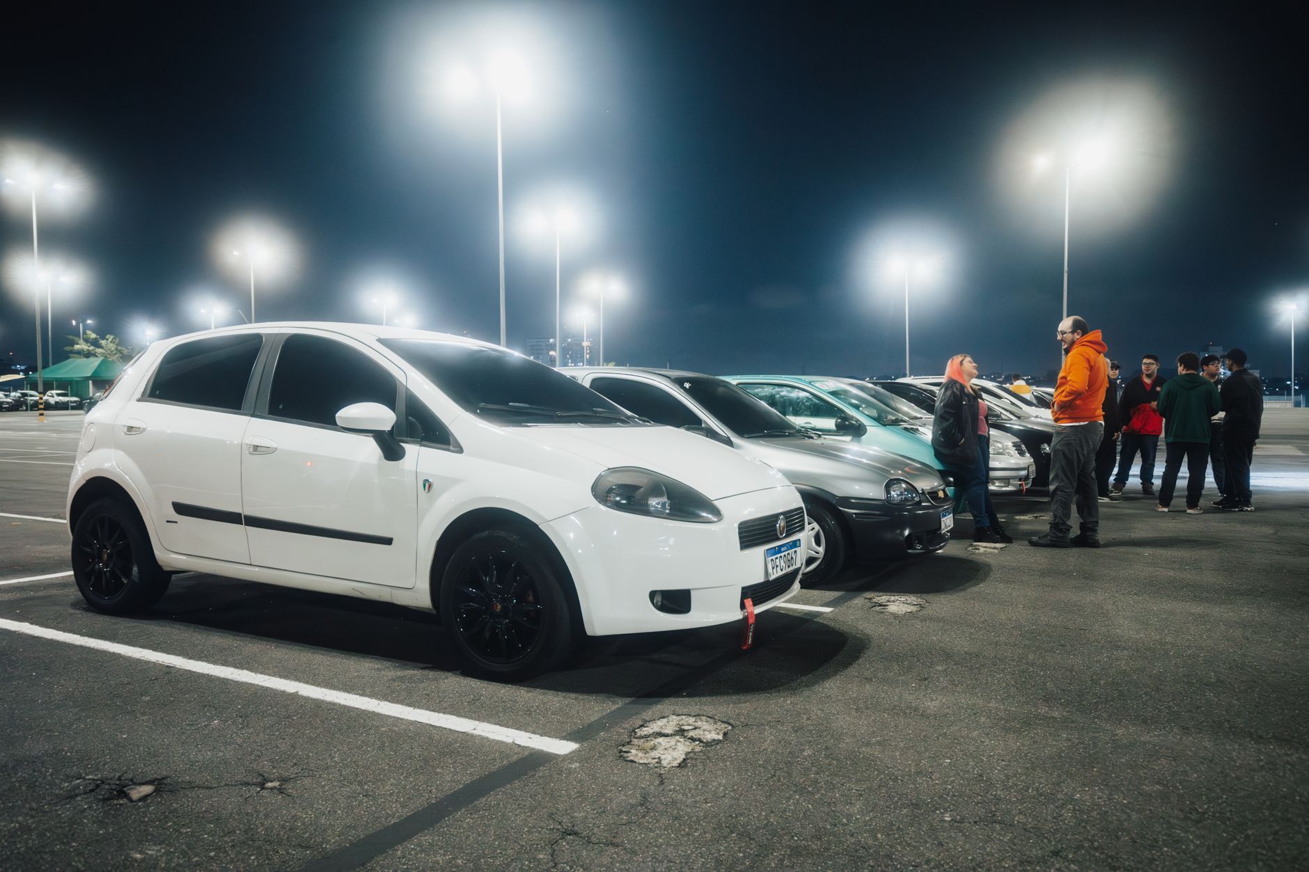 Group of parked cars and people gathering under streetlights in a parking lot at night.