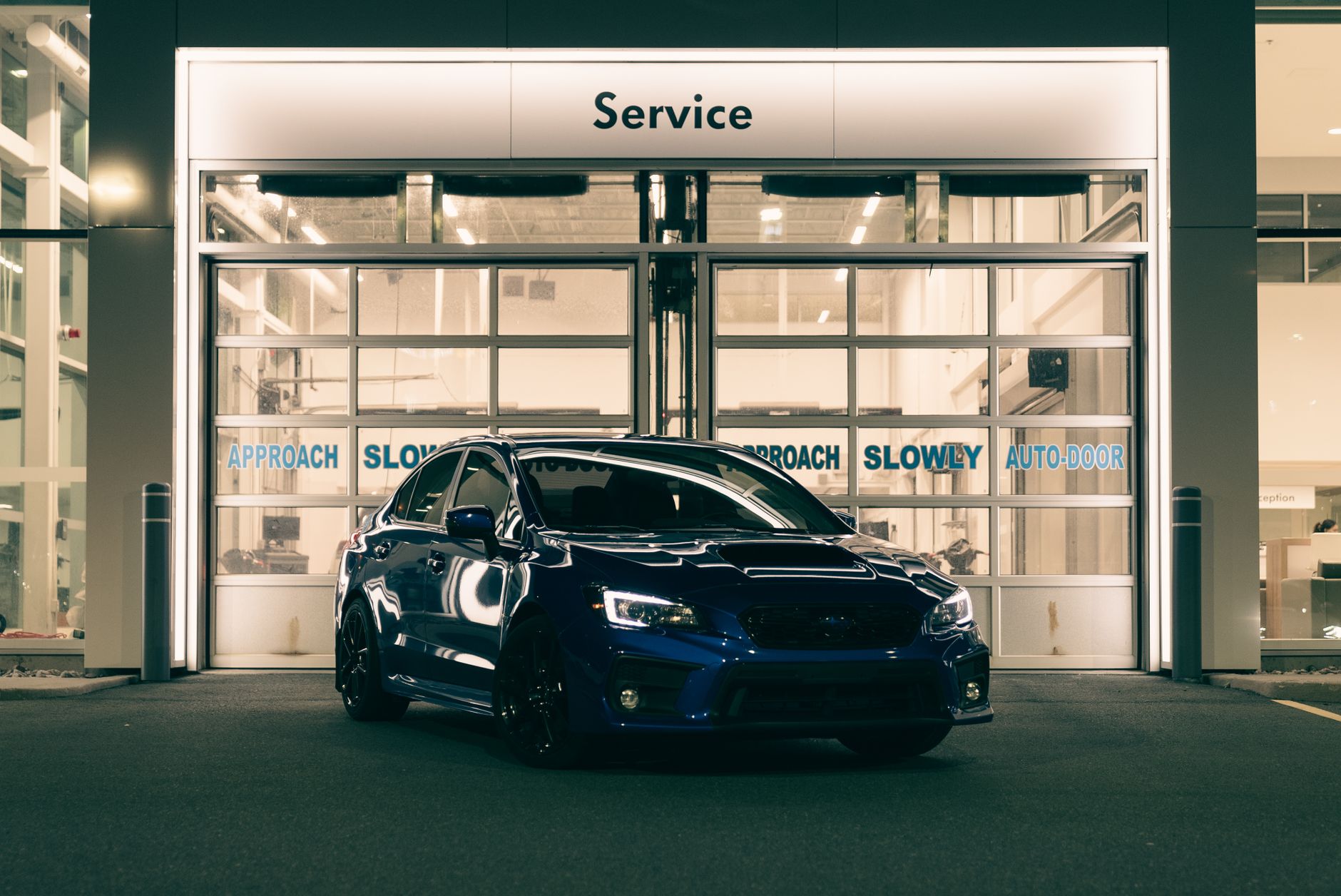 A blue sports car parked at an illuminated service center entrance during nighttime.