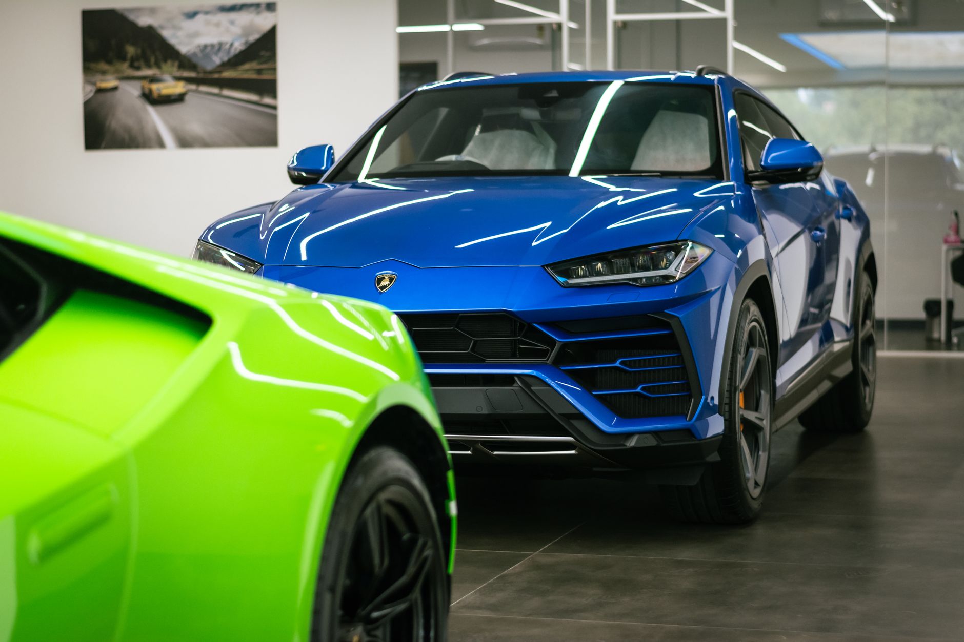 Blue Lamborghini SUV displayed in a sleek, modern showroom with a vibrant green car in the foreground.
