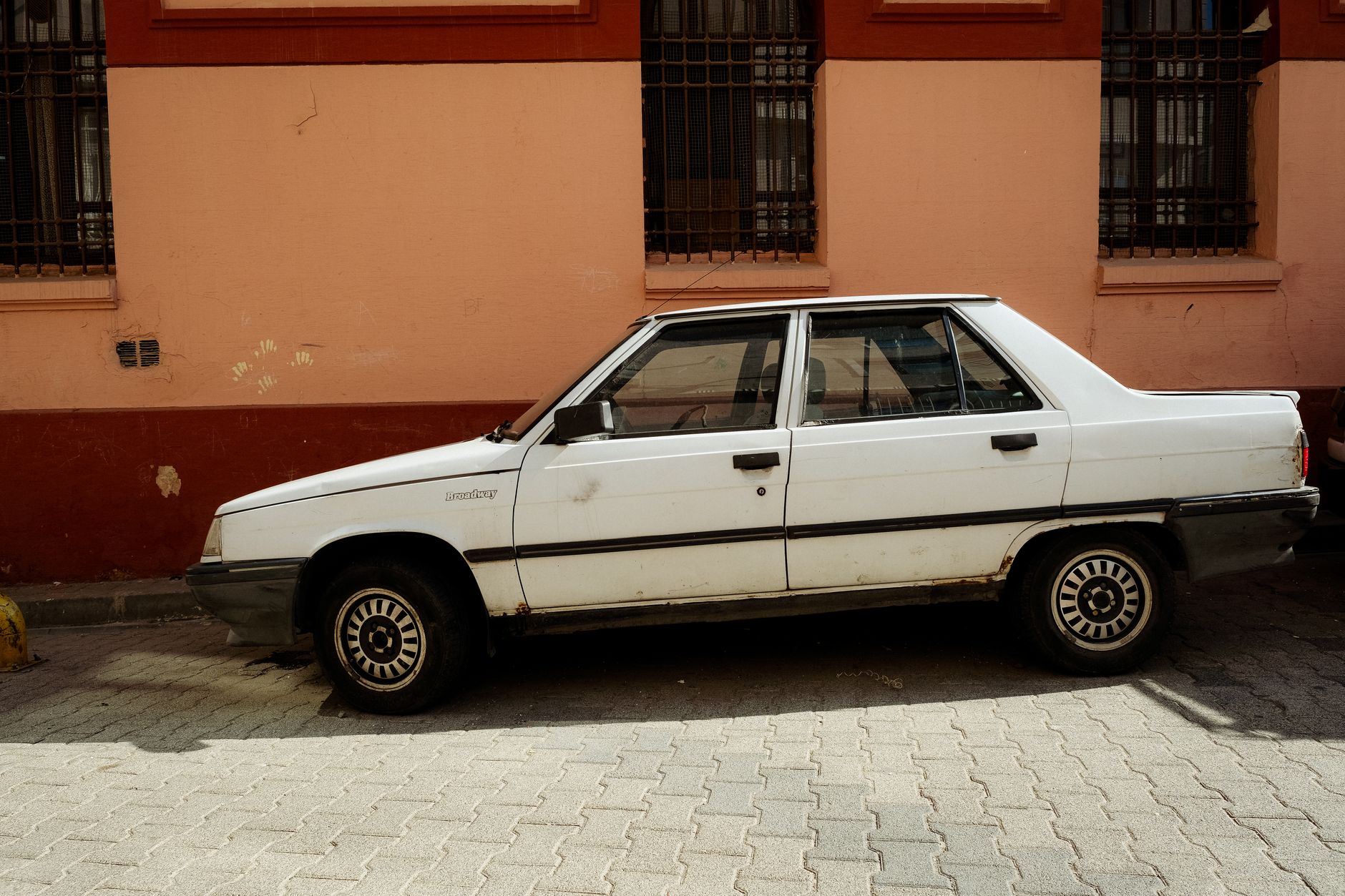 A classic white sedan parked on a street in front of a brick wall, showcasing urban decay.