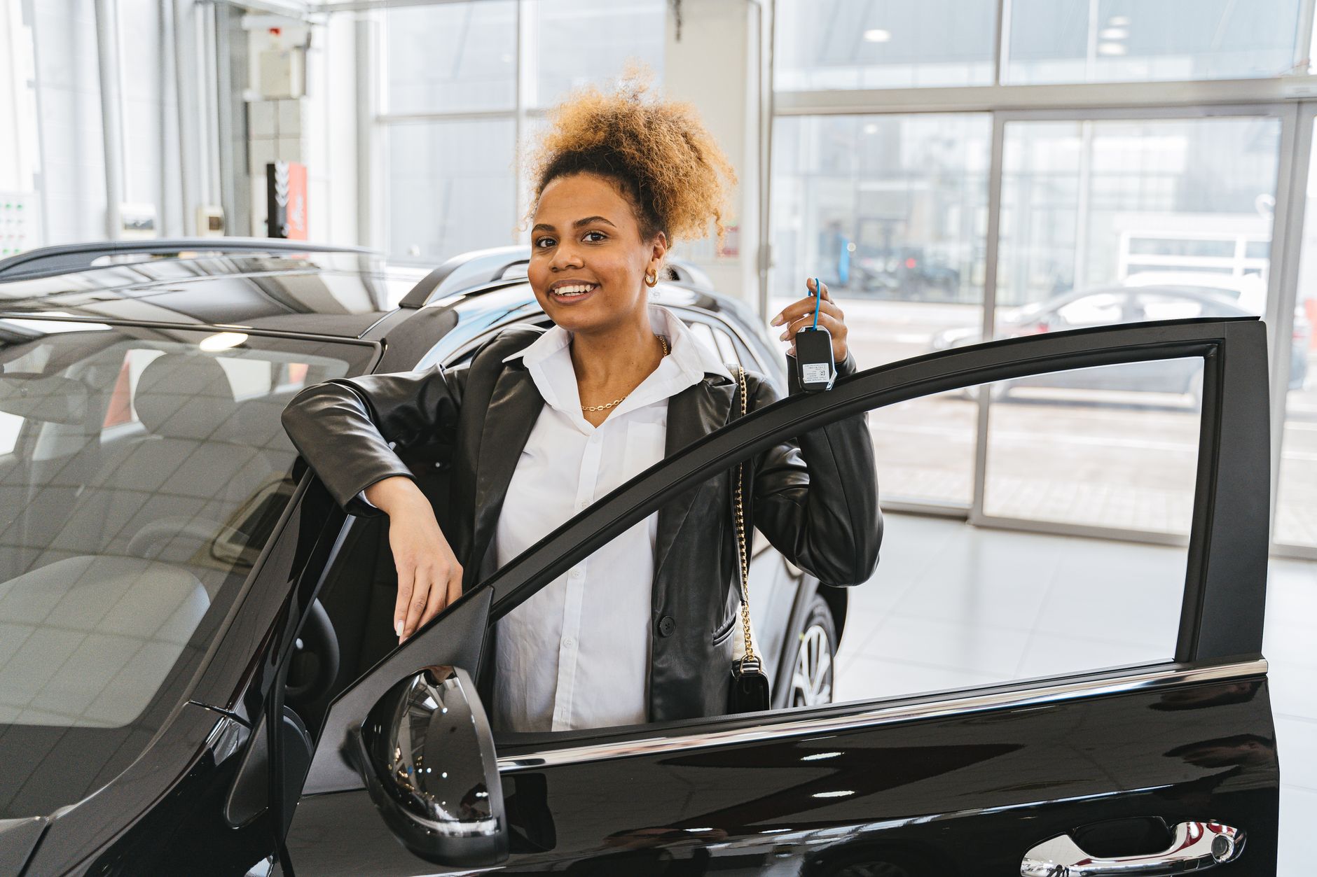 Smiling woman holding car key in dealership showroom next to new vehicle.