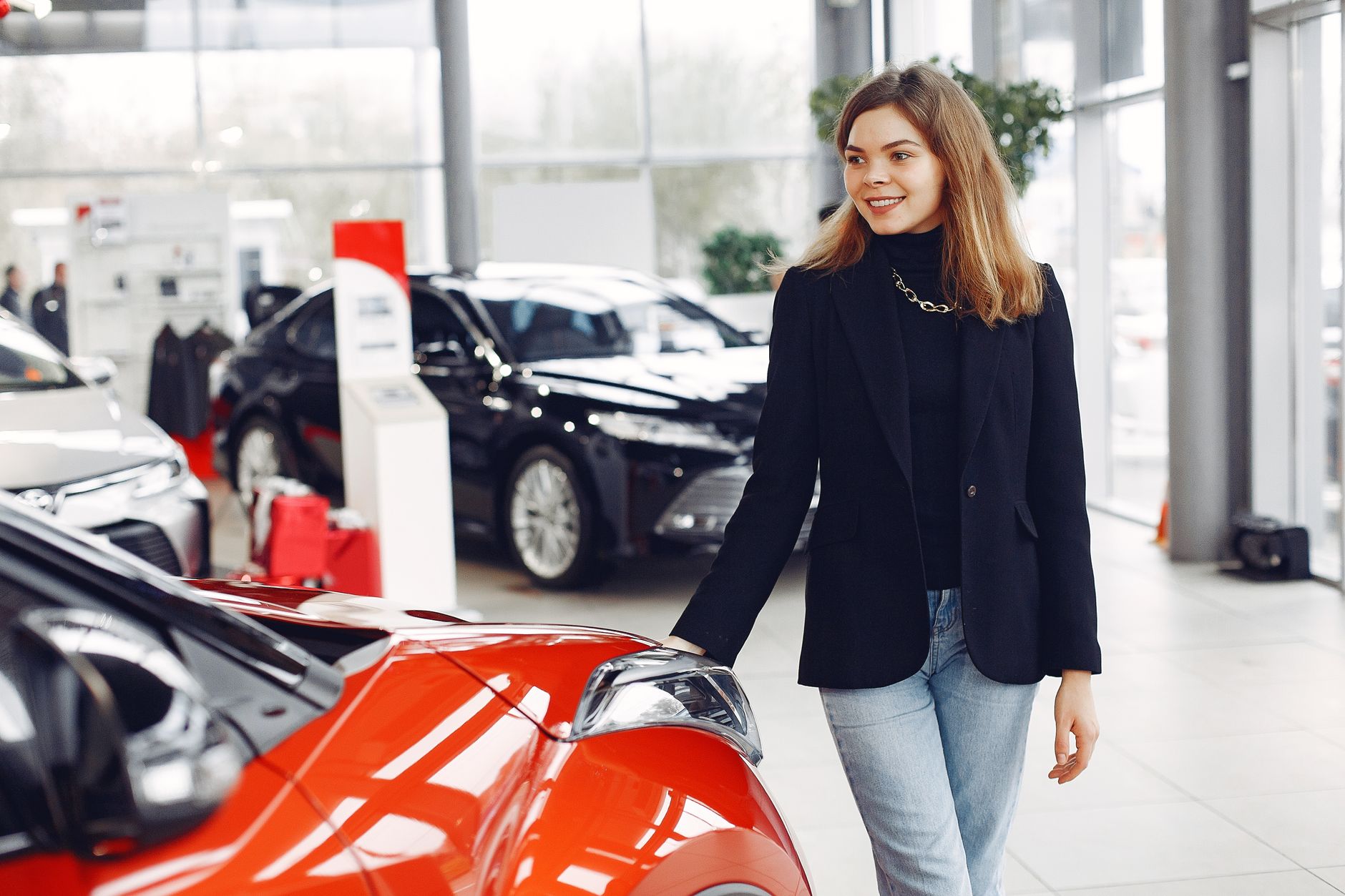 Happy stylish young female in jeans and black jacket standing near modern red shiny car in contemporary car showroom and looking at purchase dreamily