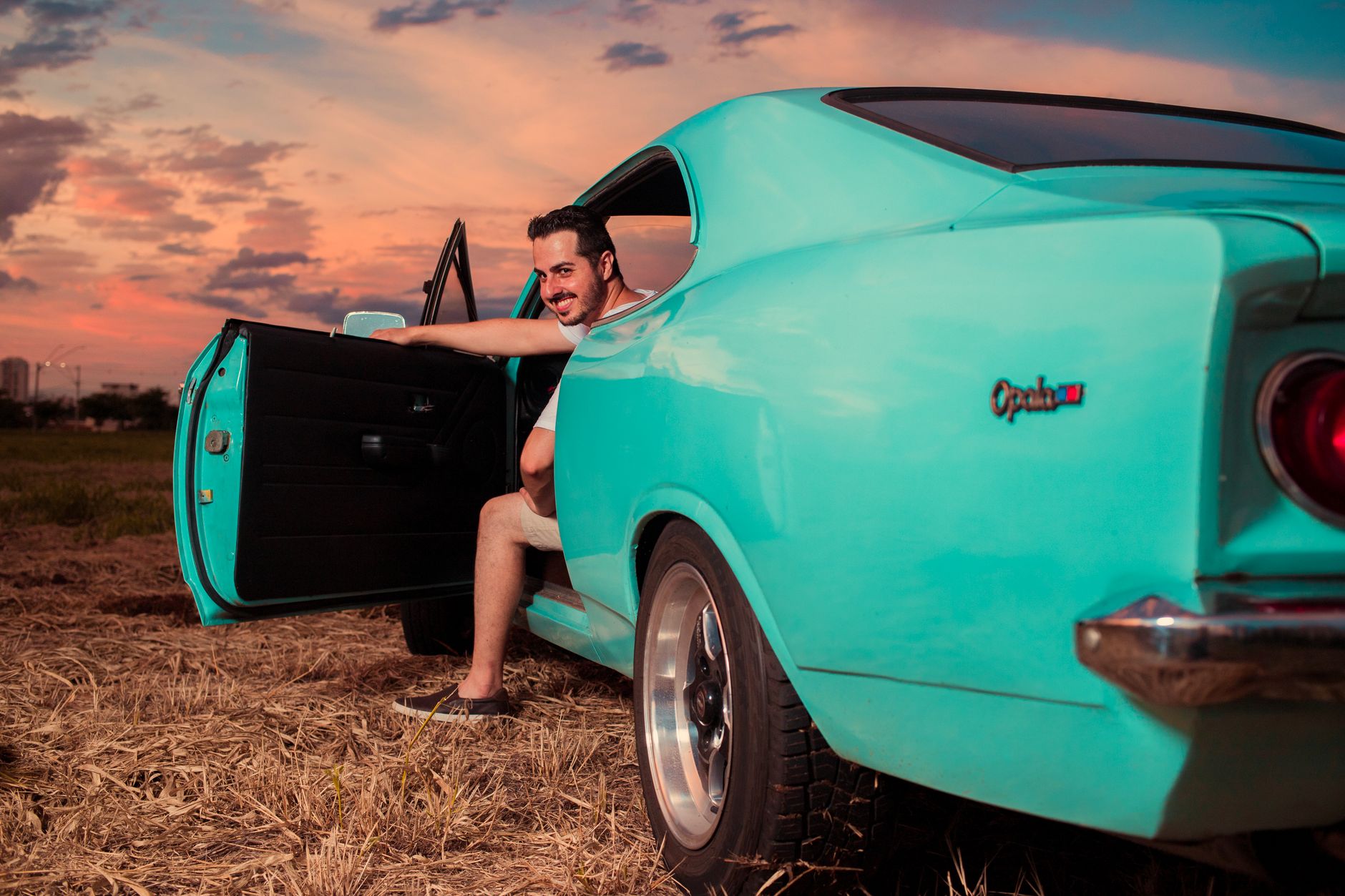 A joyful man exits a vintage teal car under a vibrant sunset sky, capturing a moment of freedom.
