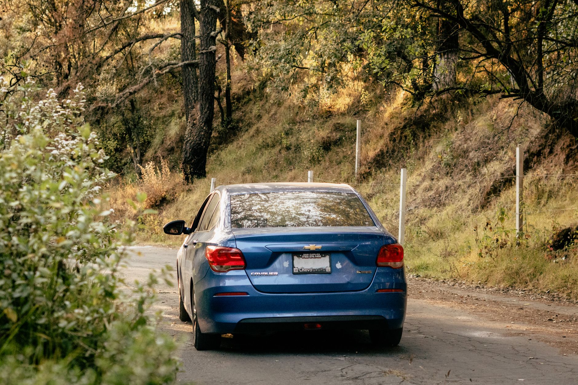 A blue Chevrolet Cavalier drives through a serene forest road in Santa María Atarasquillo, México.