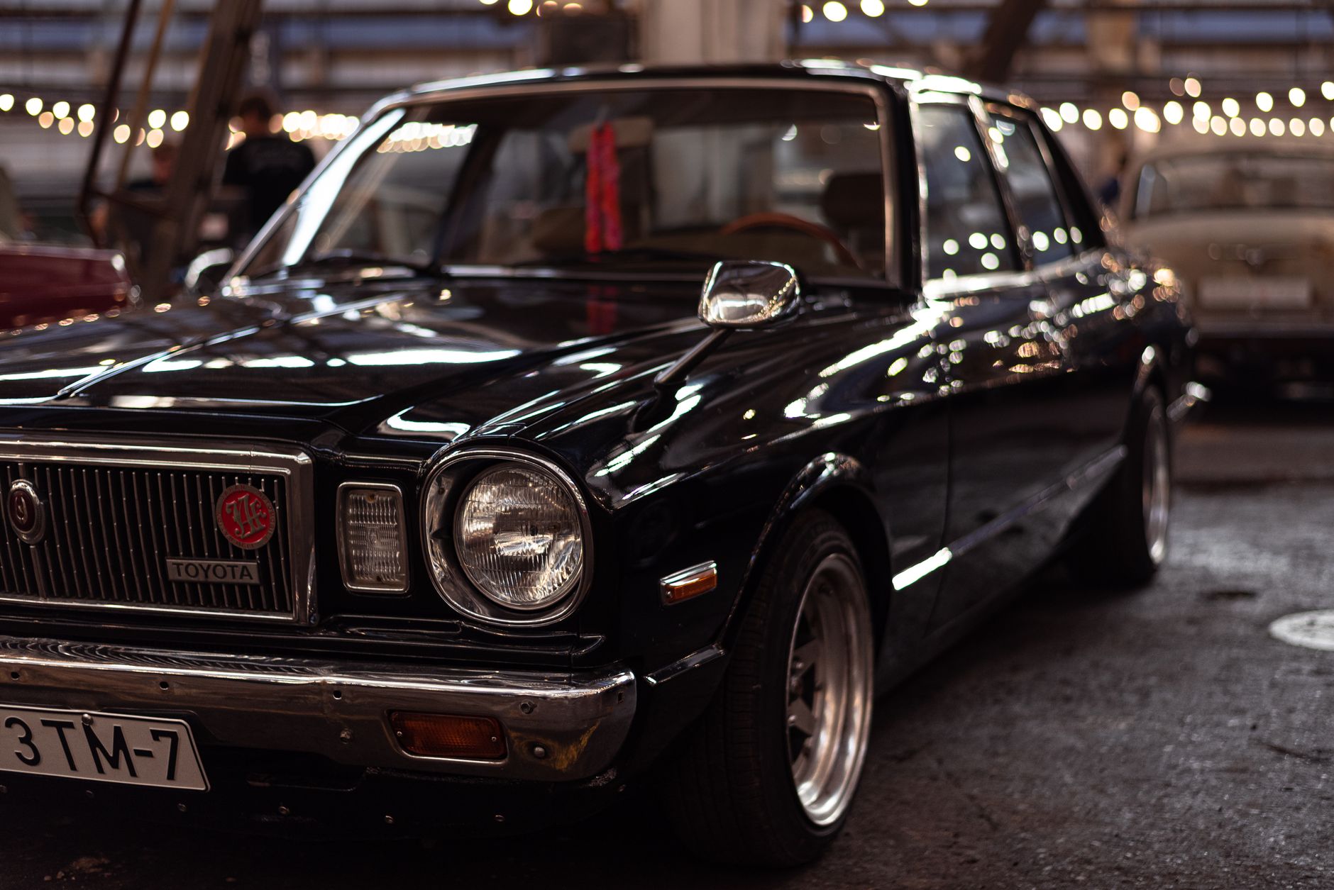 A vintage black Toyota car displayed at an indoor car exhibition with string lights.