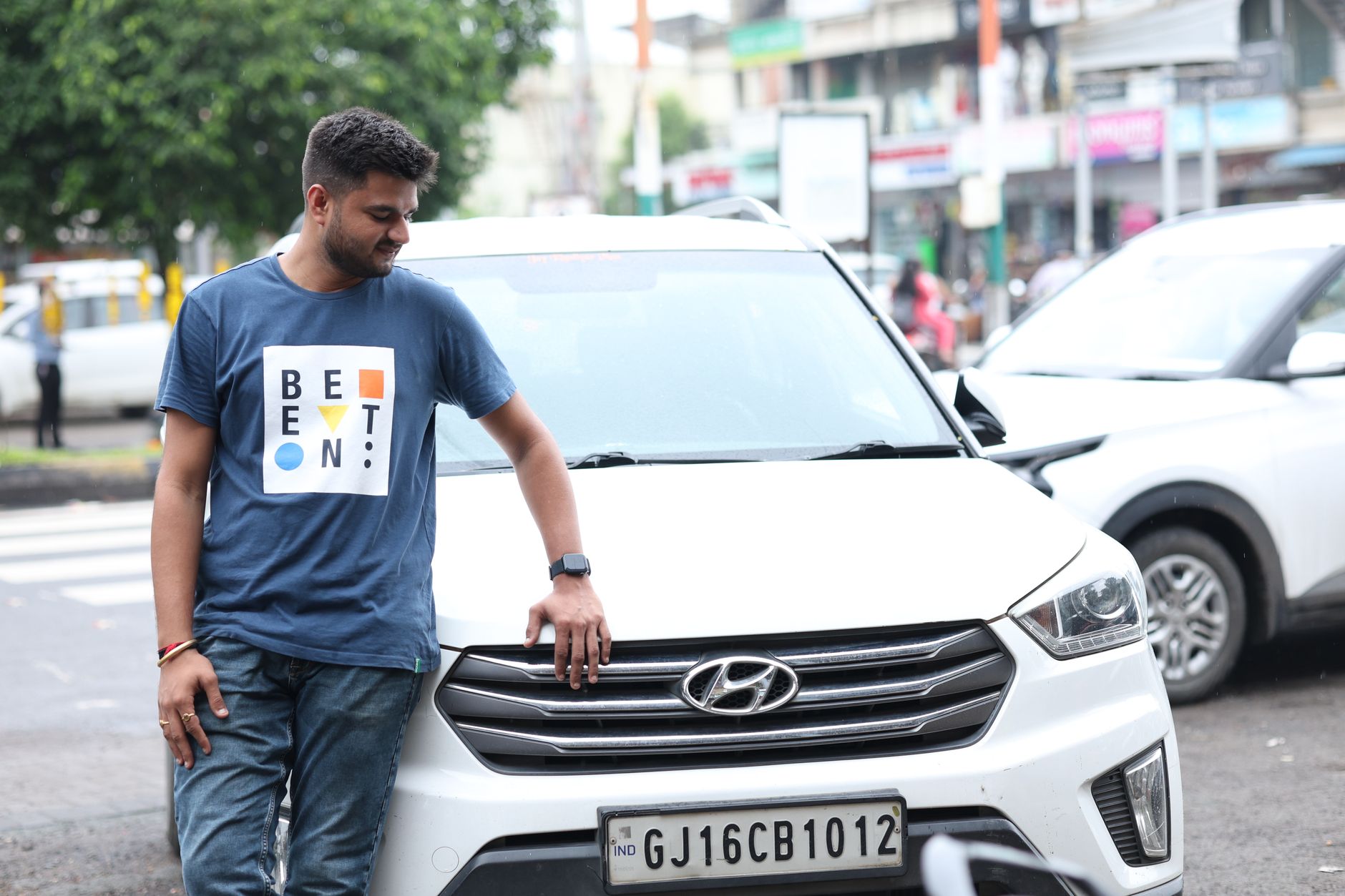 A man in a blue t-shirt casually leans on a white Hyundai car parked outdoors in an urban setting.