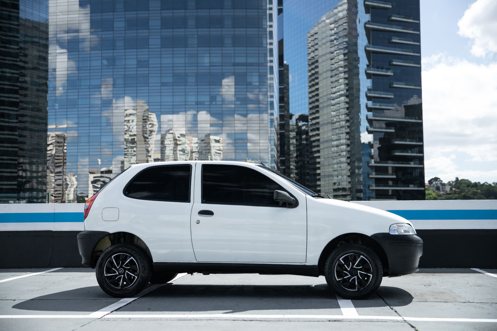 Side view of a white hatchback car parked against a backdrop of São Paulo skyscrapers.