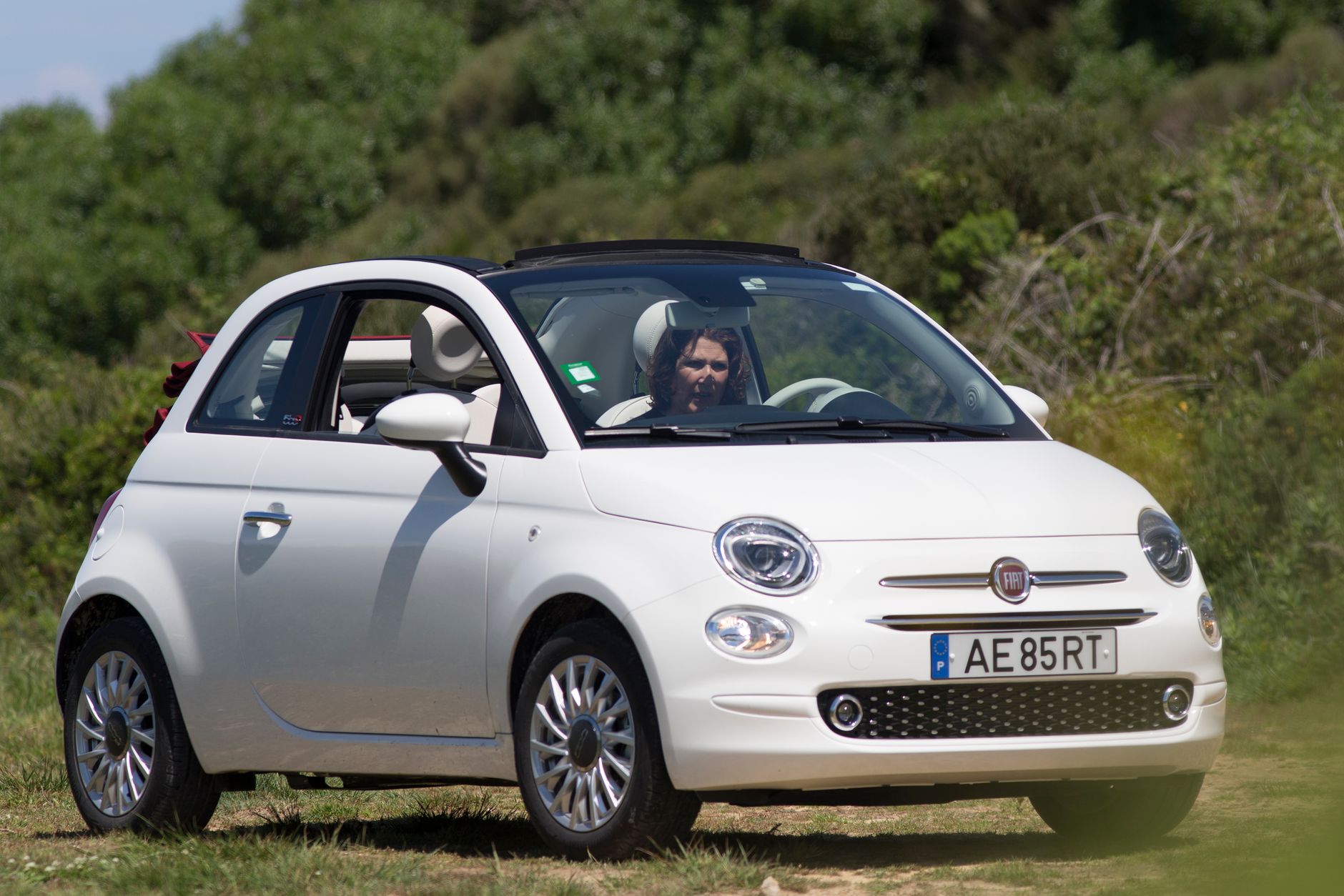 A white Fiat 500 with open roof driving on a sunny day with greenery in the background.