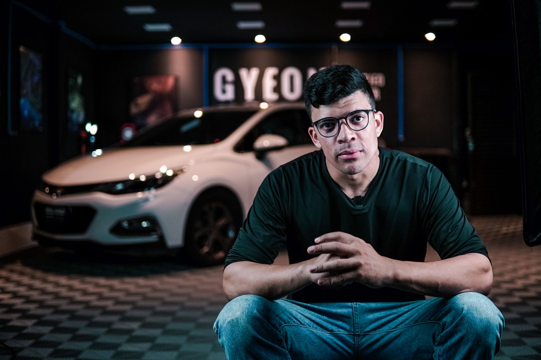 Man with eyeglasses sits in front of a white car in a showroom, hands together.
