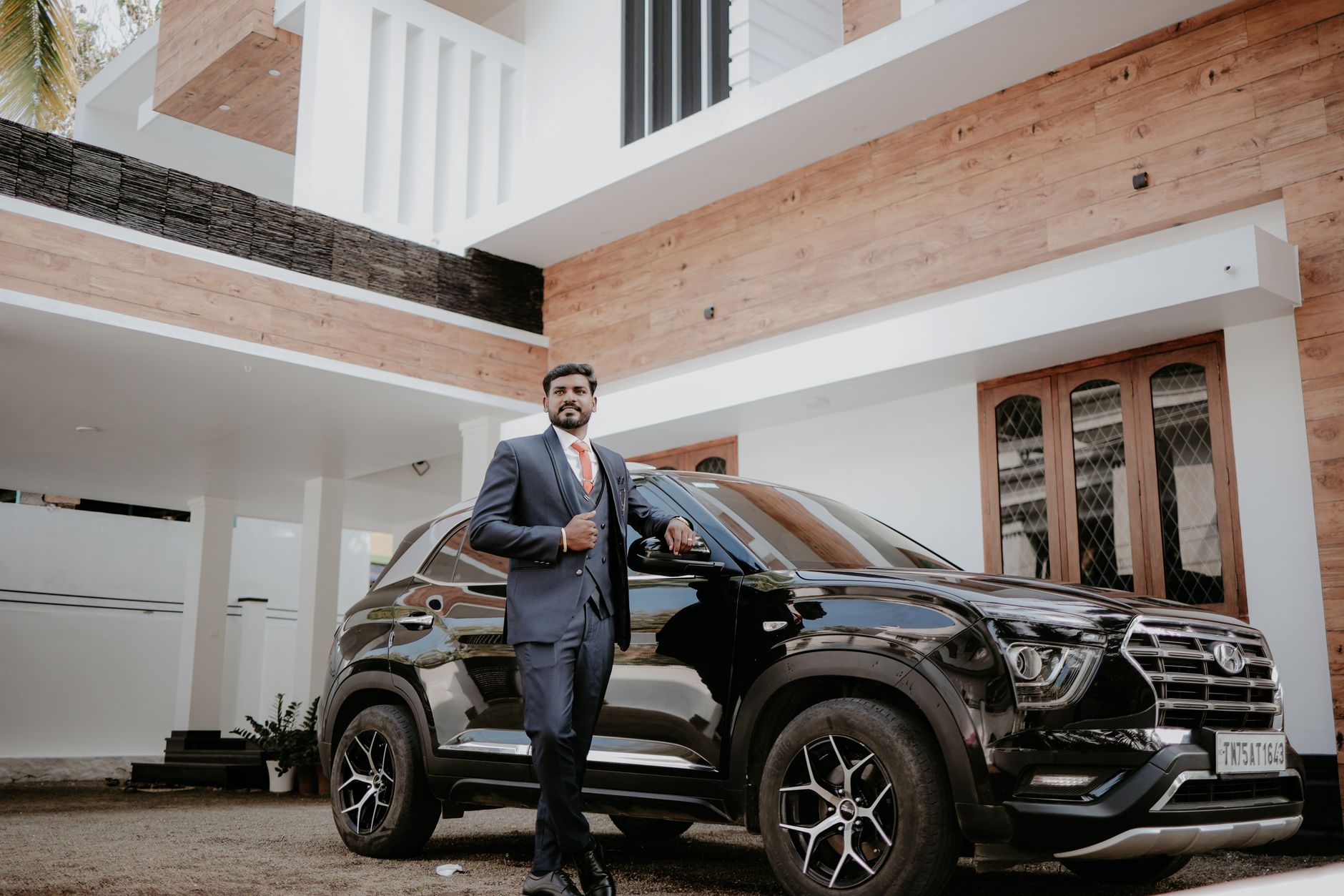 Businessman in a blue suit standing confidently next to a modern black SUV outside a stylish home in India.