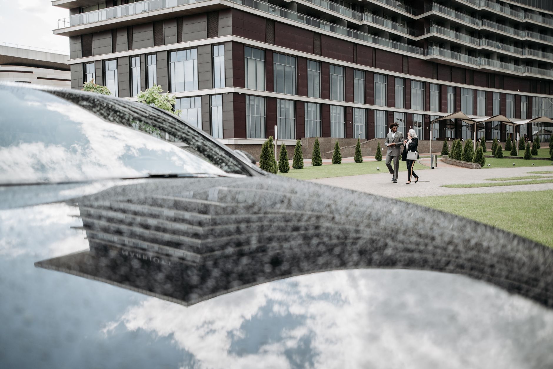 Two adults in business attire walk past a modern building, reflected in a car windshield.