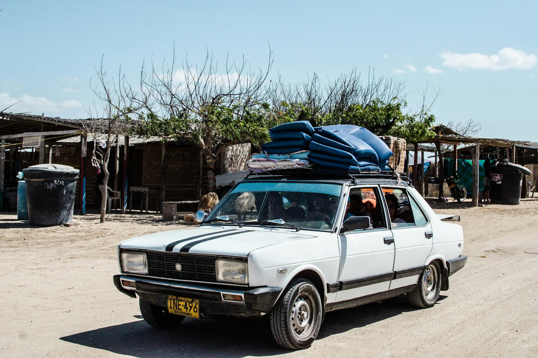 White vintage sedan with luggage on roof rack parked in a rustic rural area on a sunny day.