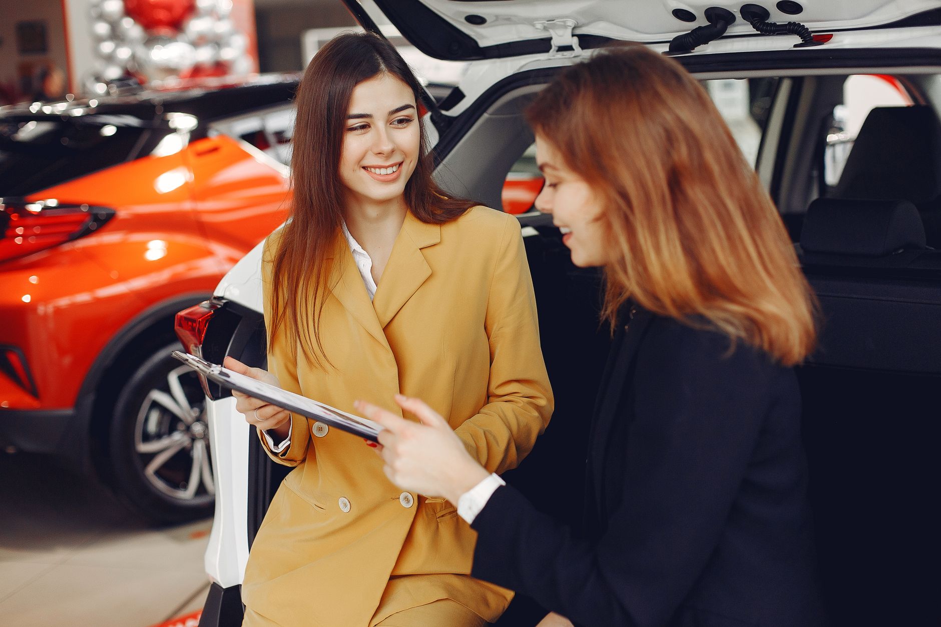 Positive smiling agent in trendy formal light brown suit discussing with happy excited female customer details of contract while leaning on car in showroom