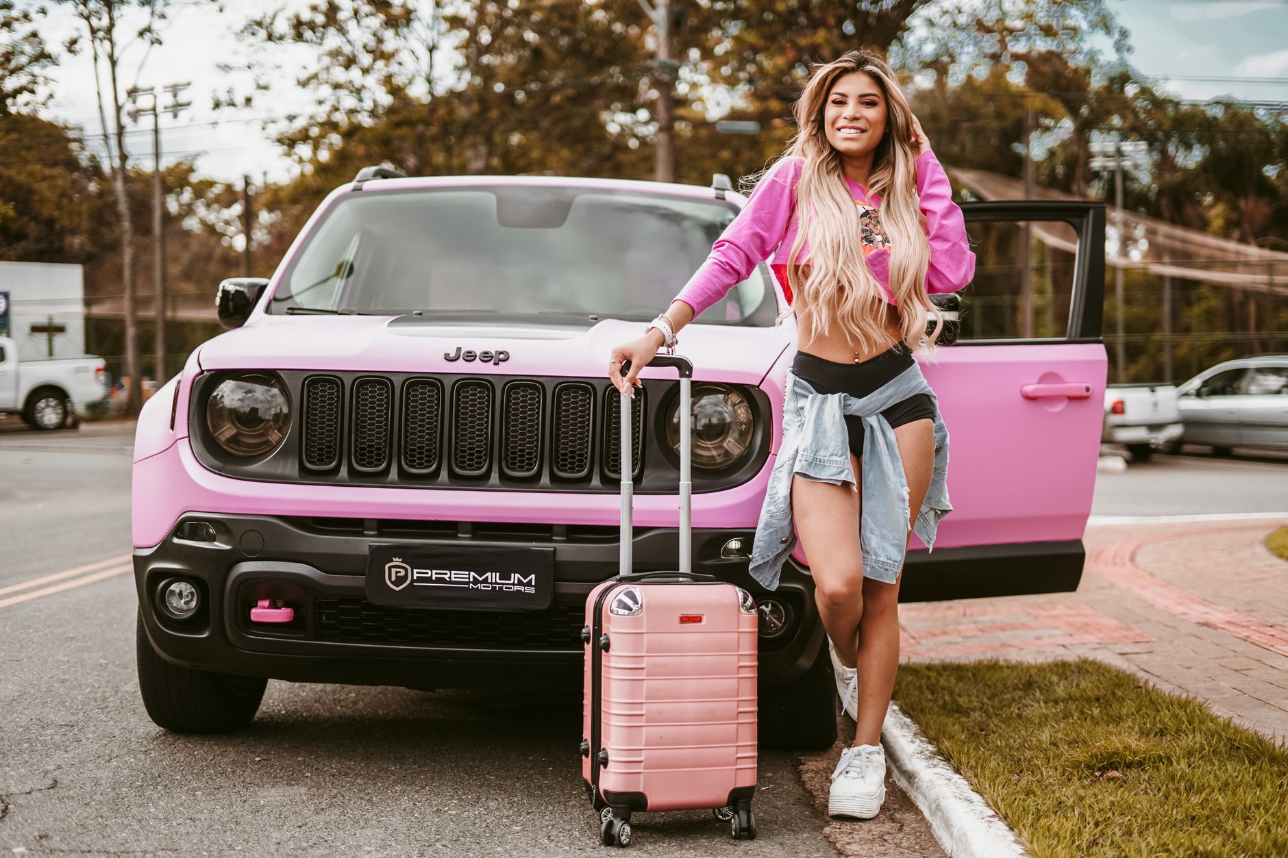 Fashionable young woman with pink top and suitcase standing by a pink SUV outdoors.