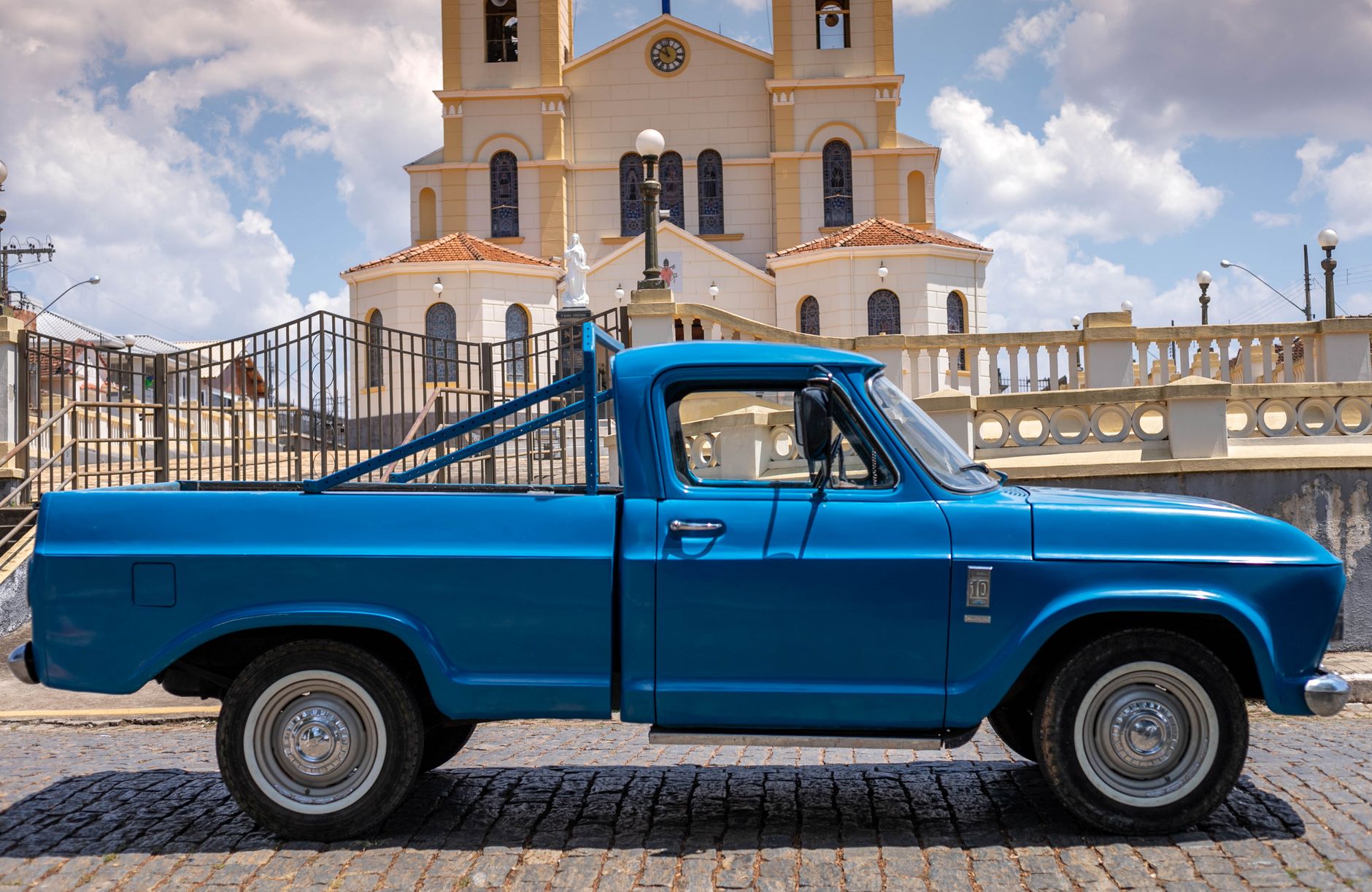 Side view of a vintage blue truck parked on cobblestone street in front of a church in Conceição do Rio Verde, Brazil.